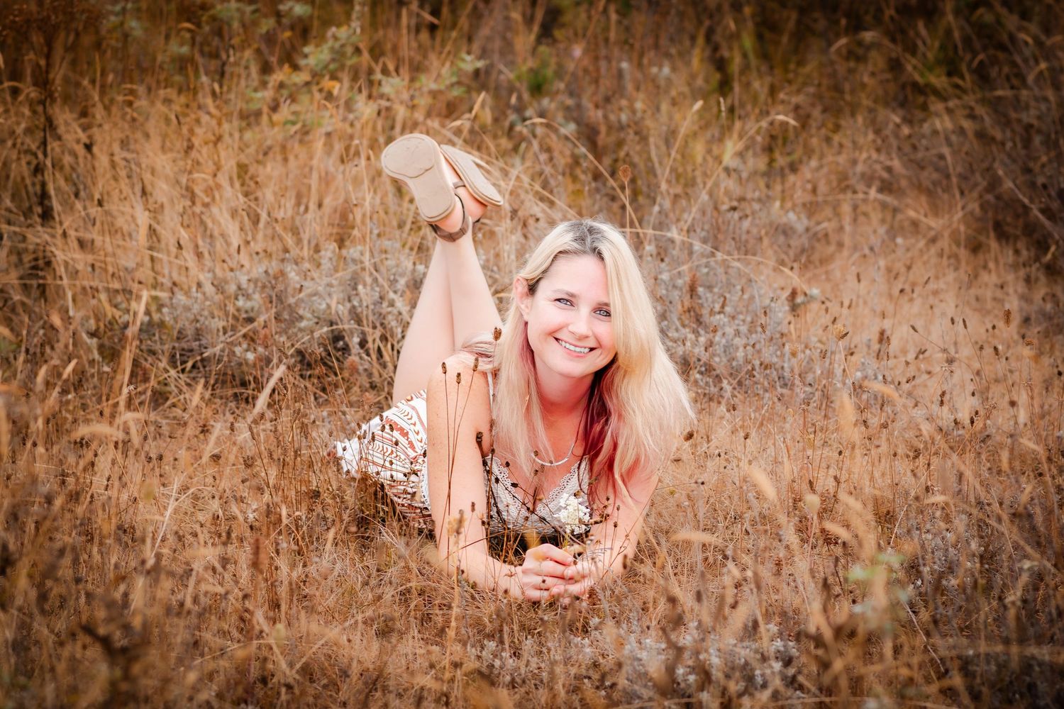 Smiling blonde woman lying in dry golden grass field wearing floral dress with feet up in casual outdoor portrait.