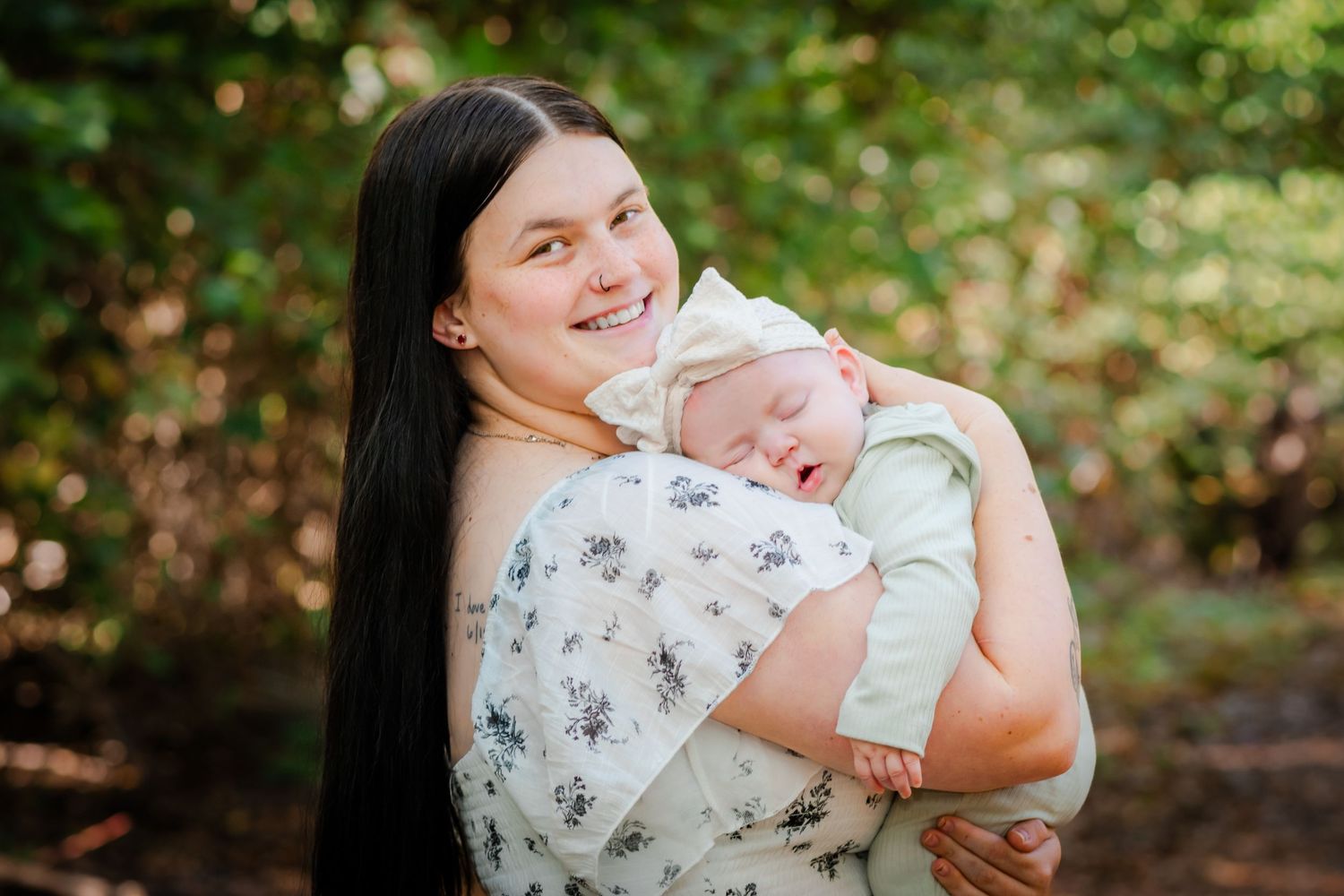 Smiling young mother with long dark hair holds sleeping newborn baby outdoors in green nature setting.