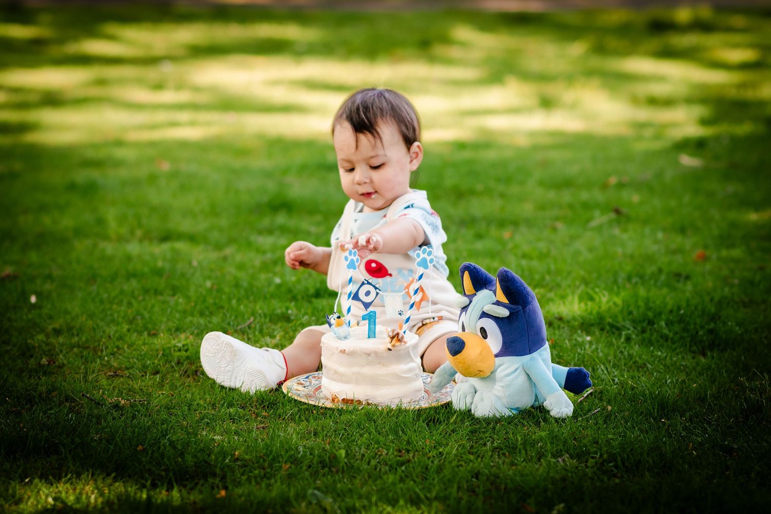 Baby sits on grass playing with toys and stuffed animals in dappled sunlight.