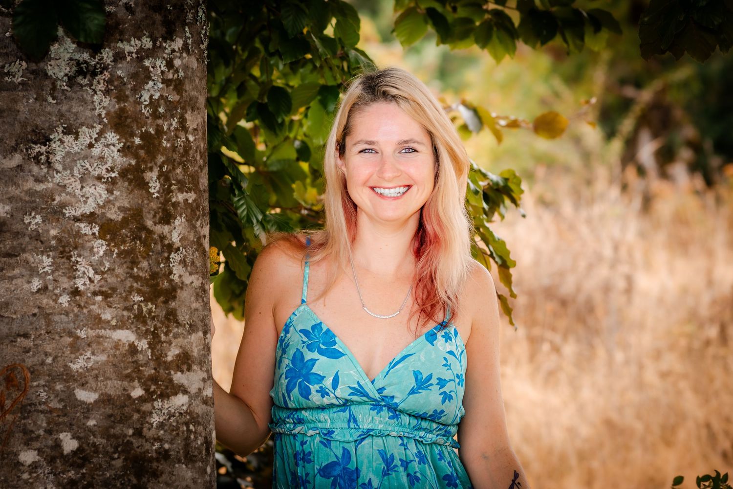 Smiling blonde woman in blue floral dress leaning against a tree in a sunny outdoor setting.