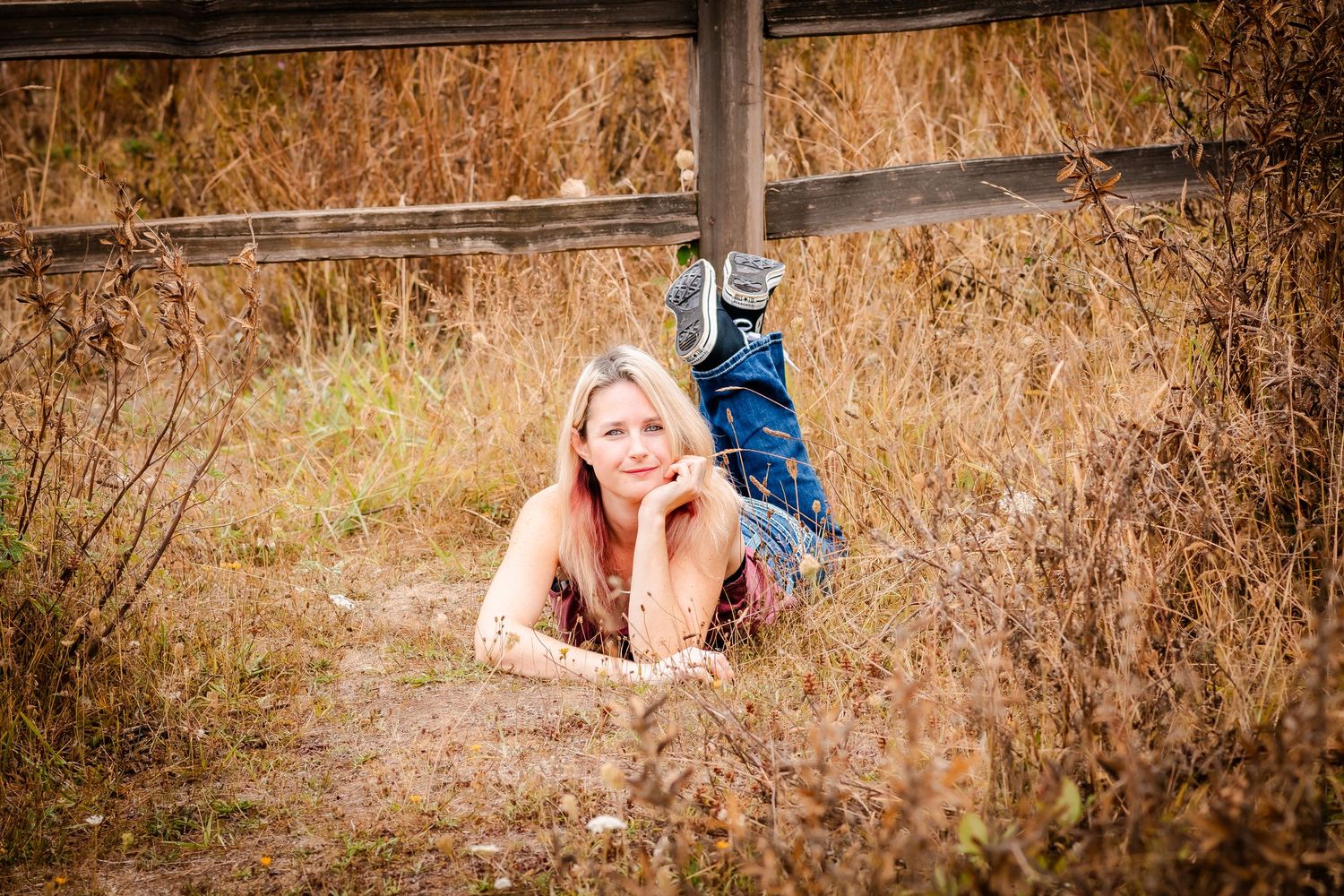 A young blonde woman in jeans lies in dry golden grass near a wooden fence, smiling outdoors.