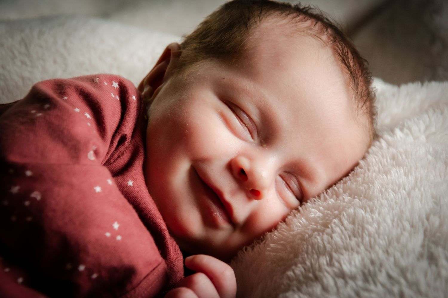 Sleeping newborn wearing burgundy outfit on cream fur backdrop.