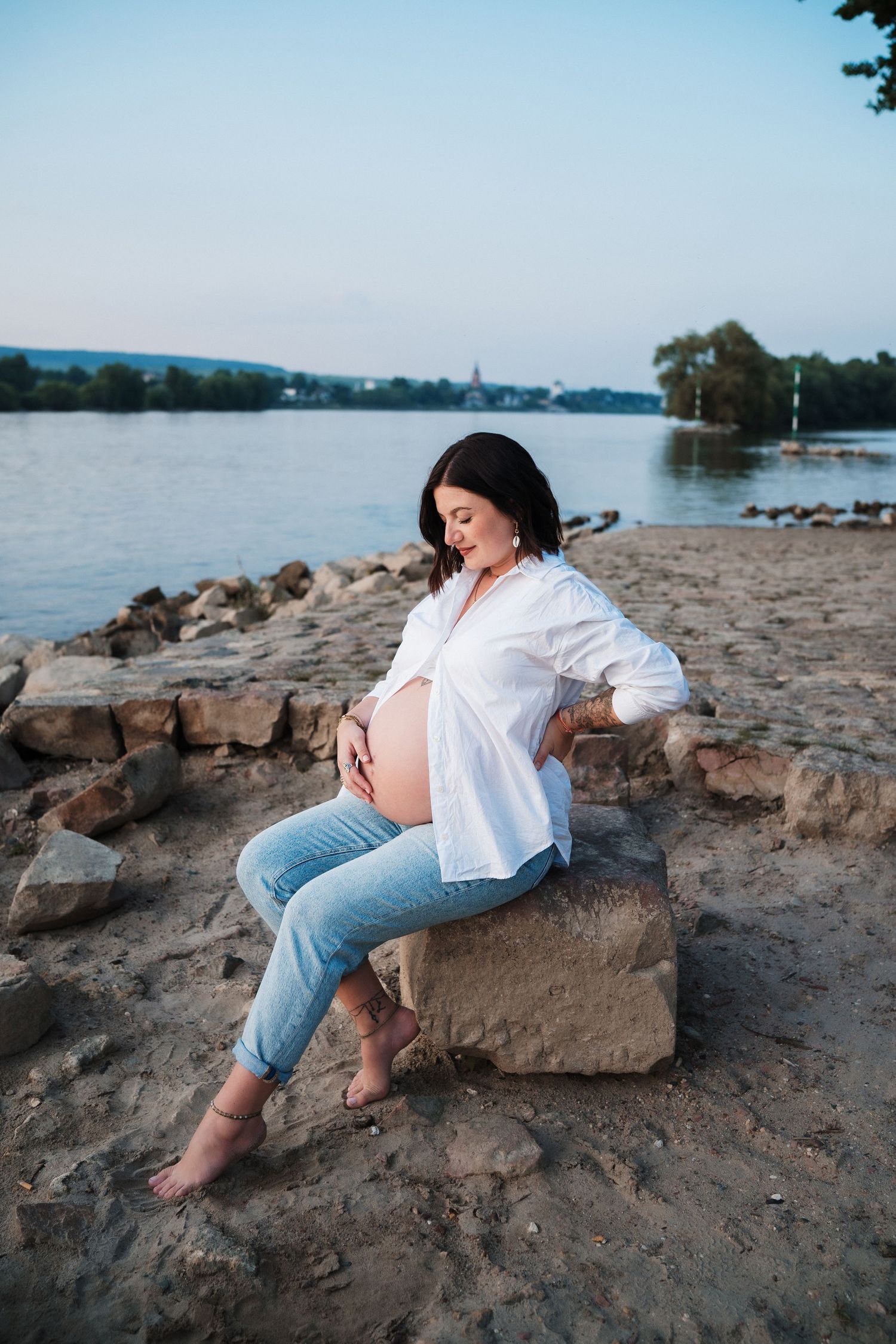 Expectant mother in white shirt and jeans sits on rocks by a serene lakeside.