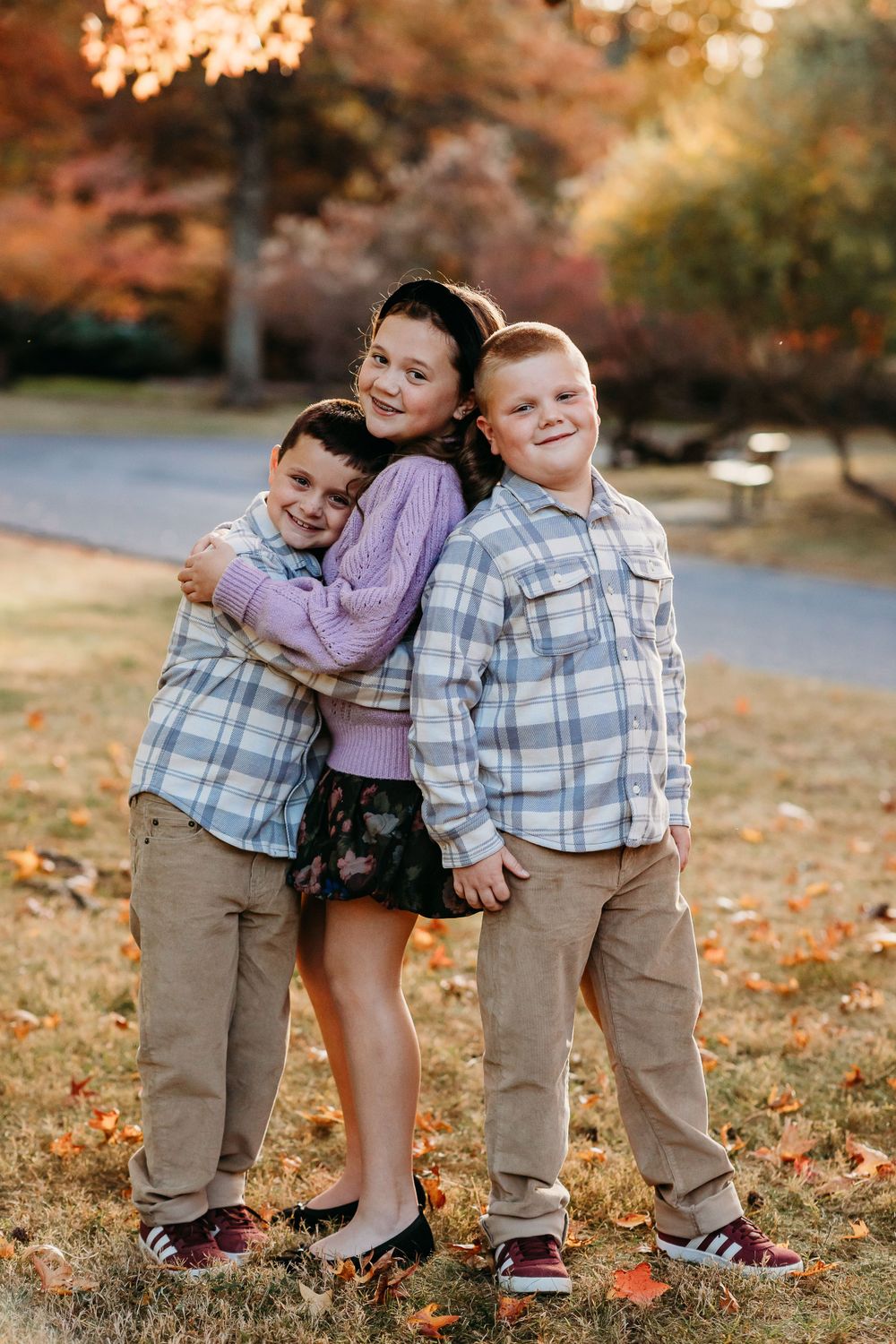 Three siblings smile and pose together on an autumn day with fall foliage creating a warm golden backdrop.