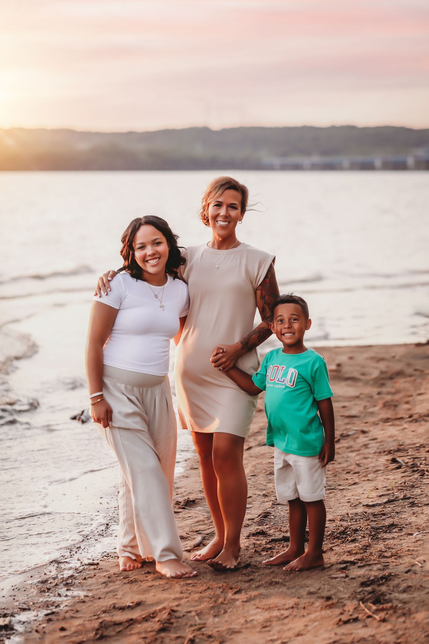 A family stands together on a sandy beach at sunset with water and mountains in the background.