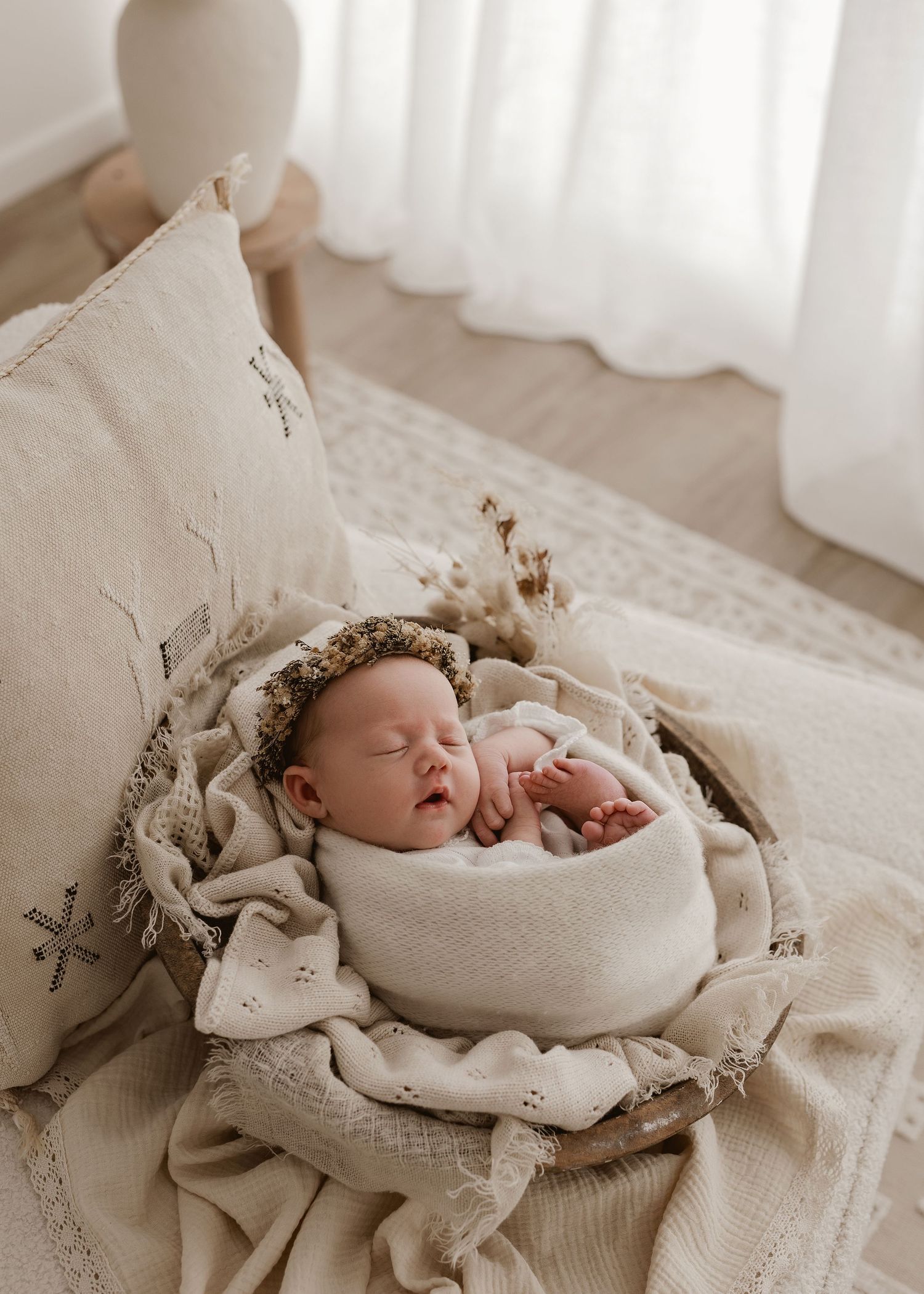 A sleeping newborn wrapped in beige swaddle blankets lies on textured fabric with a delicate floral crown.