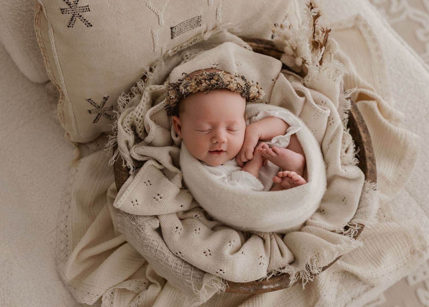 Sleeping newborn wrapped in soft beige fabric with a delicate headband, nestled in a cozy neutral-toned setting.