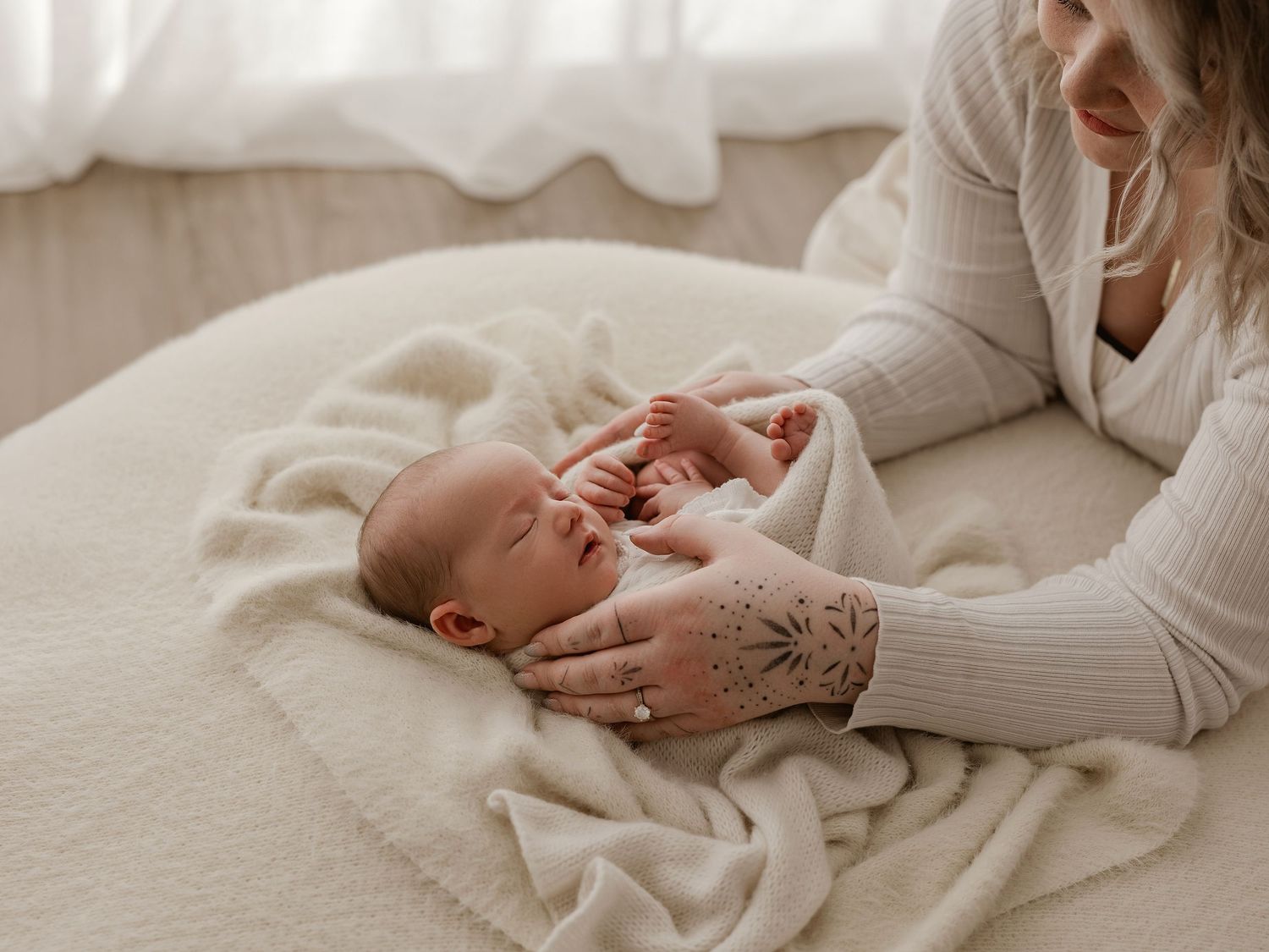 A small baby wearing a white onesie lies peacefully on a soft cream-colored blanket while being gently touched.