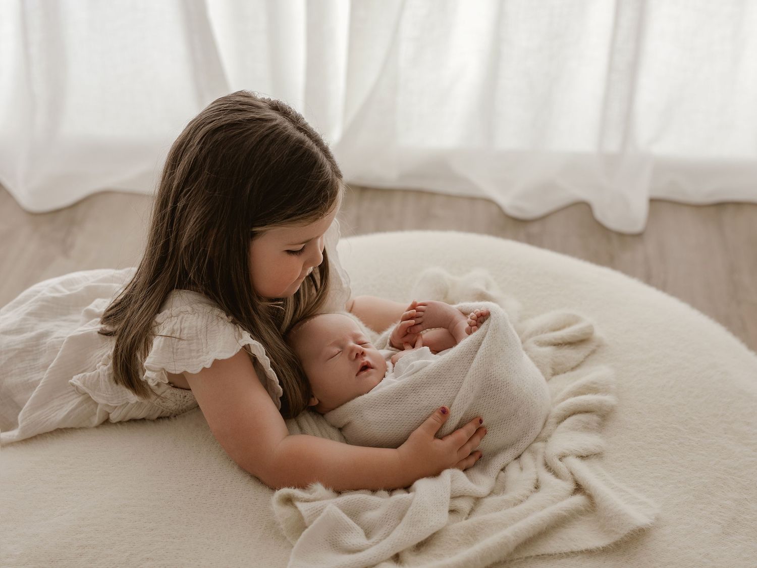 Young child in white dress gently holds newborn baby wrapped in soft blanket on light-colored floor.