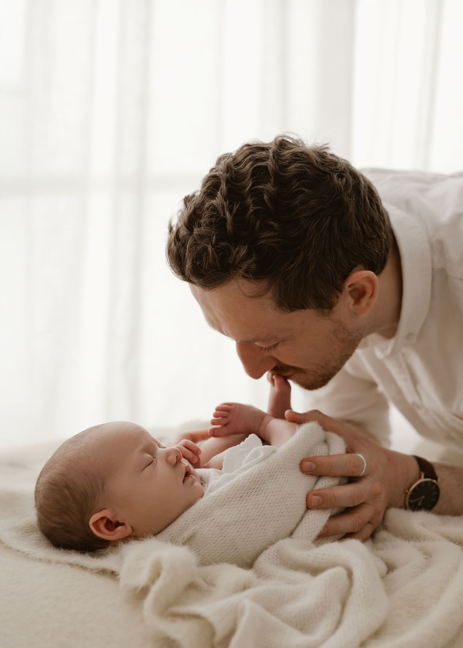 A parent in white shirt lovingly plays with and kisses a baby wearing cream colored clothing on a white bed.