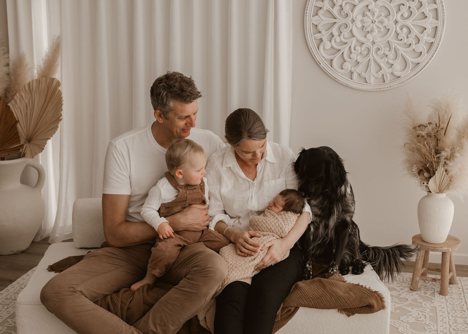 Family relaxes together on couch in neutral-toned living room with decorative wall medallions and dried flowers.
