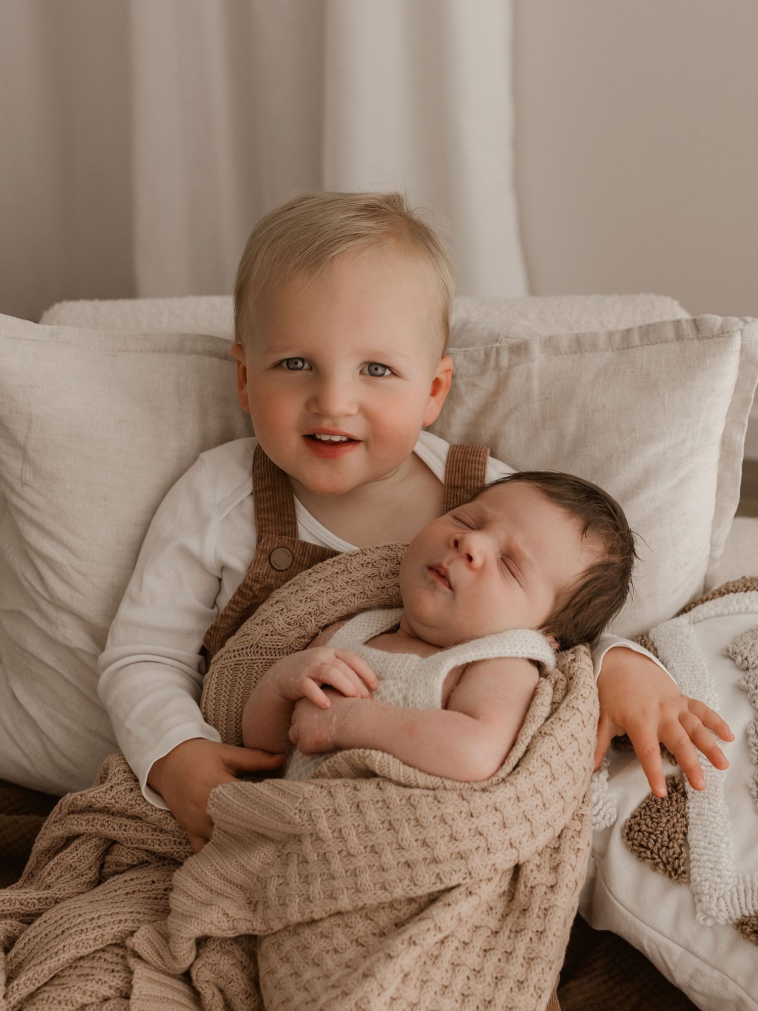Two babies snuggling together on white bedding while wearing neutral colored clothing and sharing sweet moments.