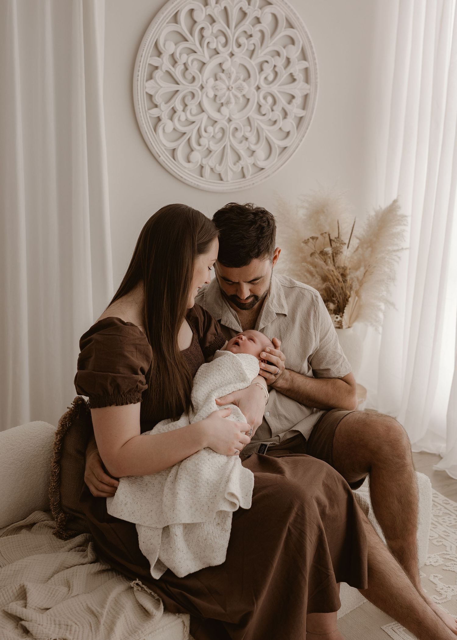 A couple shares tender moments with their newborn baby while sitting on a bed with white curtains and decor.