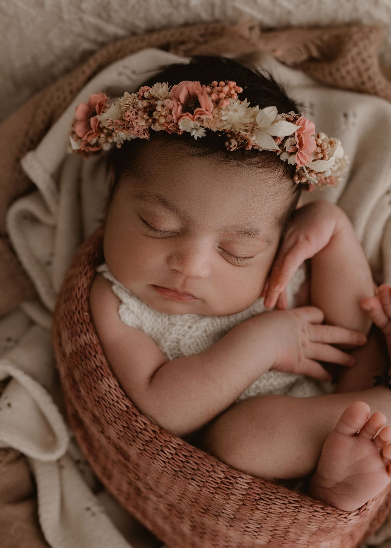 A sleeping newborn wearing a floral crown lies peacefully in a cozy pink woven basket.