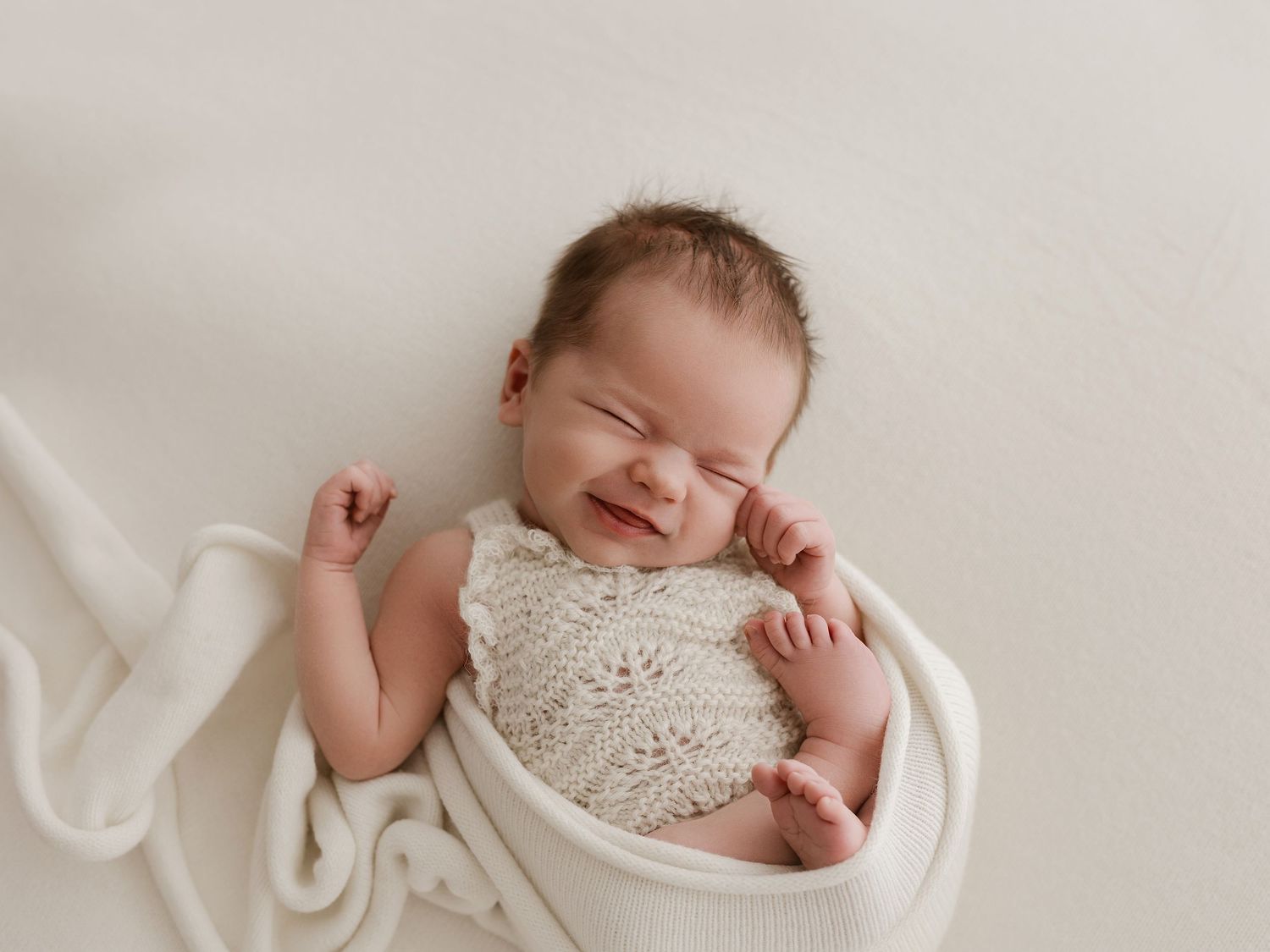 Newborn baby wrapped in white blanket smiles while wearing knit lace top during professional photo session.