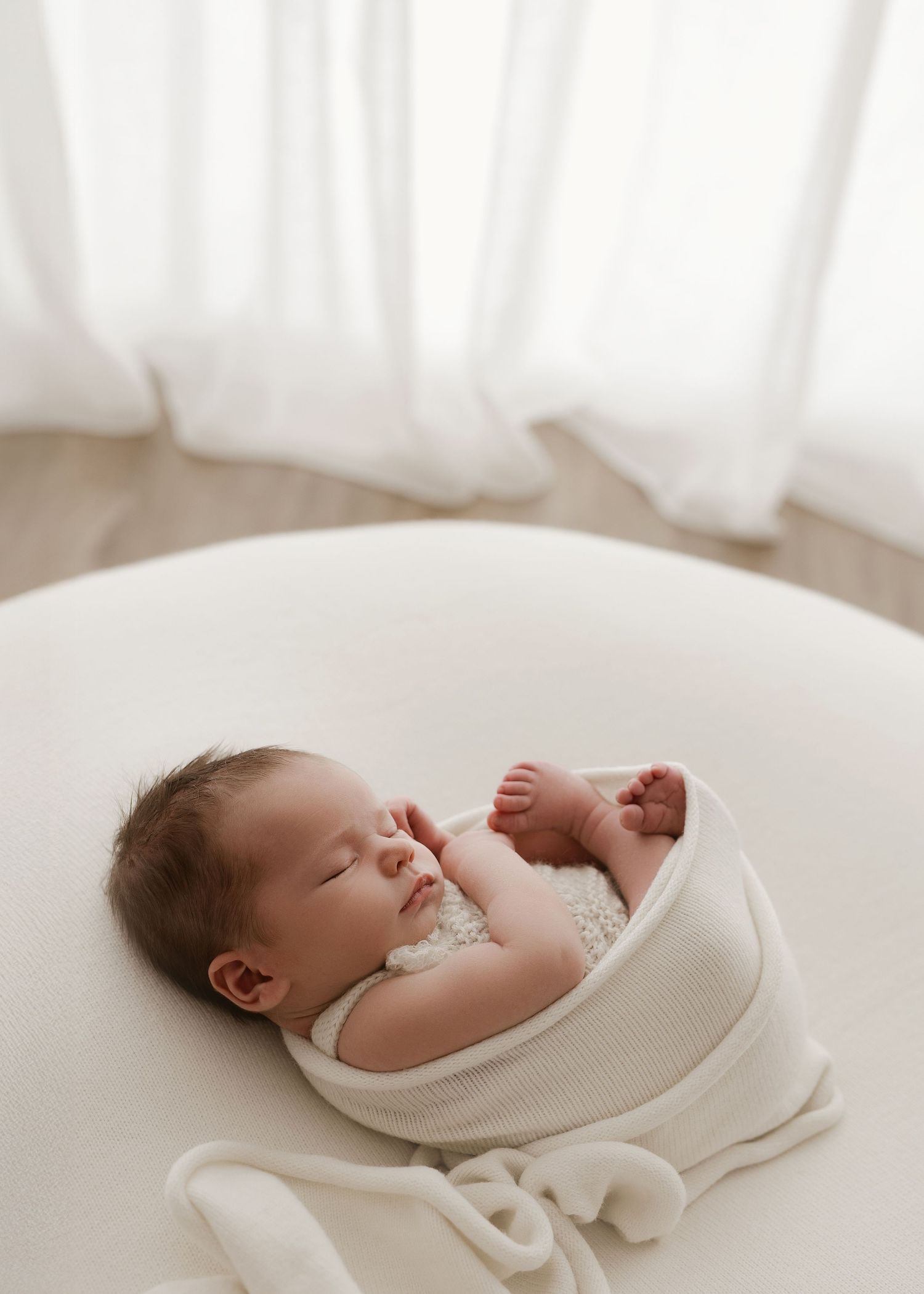 Peaceful sleeping baby wrapped in soft white swaddle blanket lying on cream colored surface in natural light.