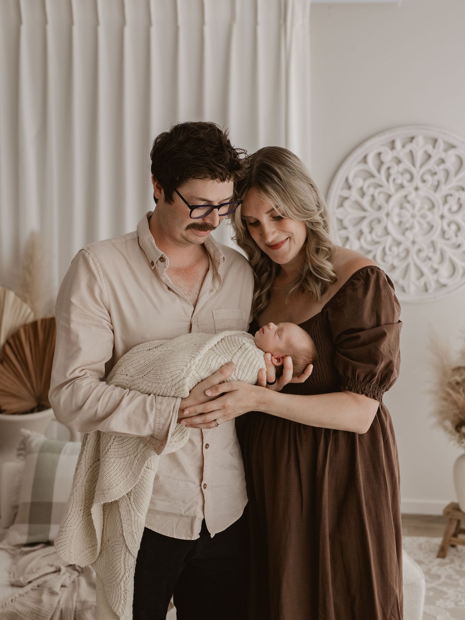 A couple in beige and brown outfits embrace tenderly in a minimalist indoor setting with white decorative wall panels.