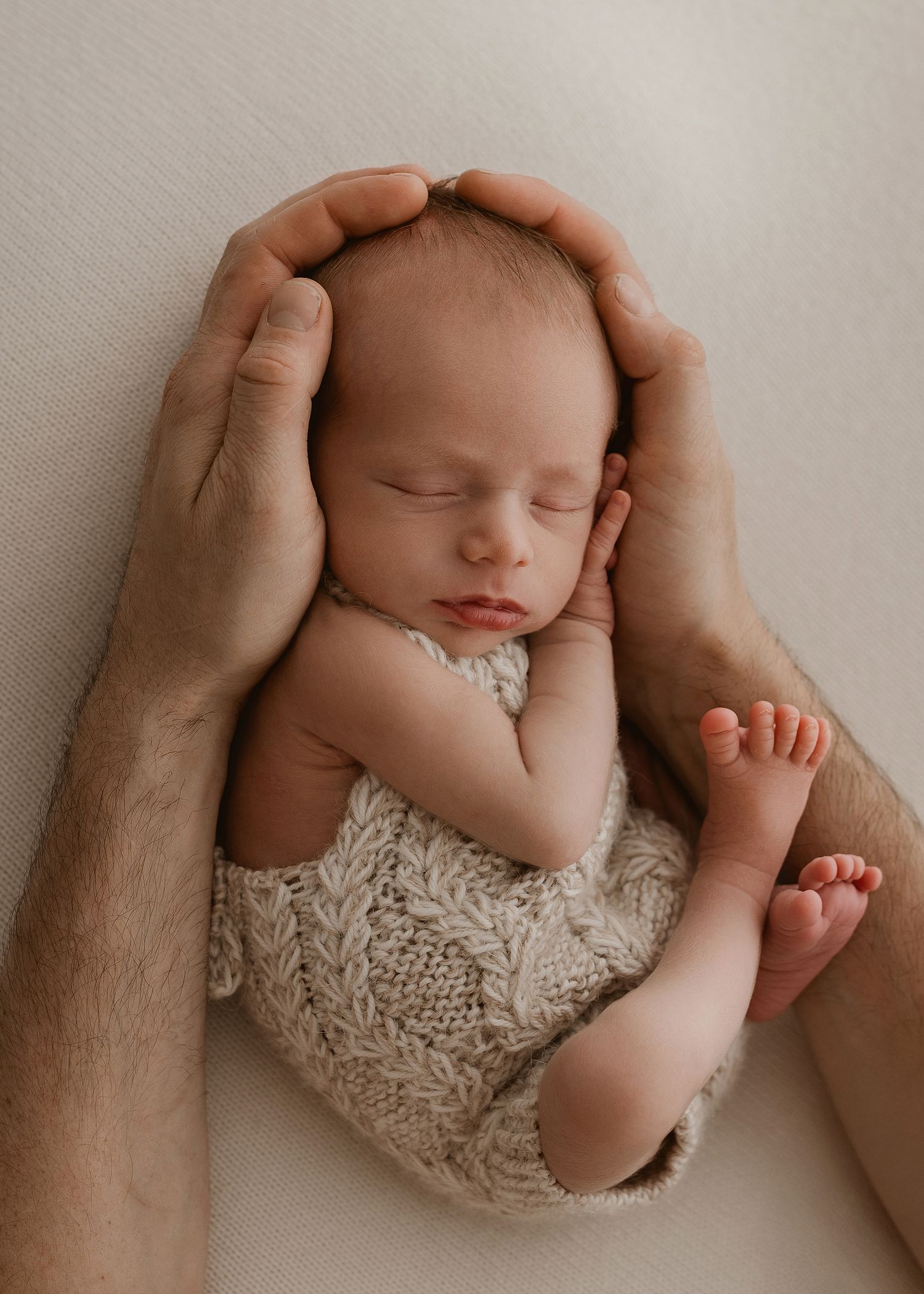 Gentle hands cradle a sleeping newborn wearing delicate knit clothing against a cream-colored background.