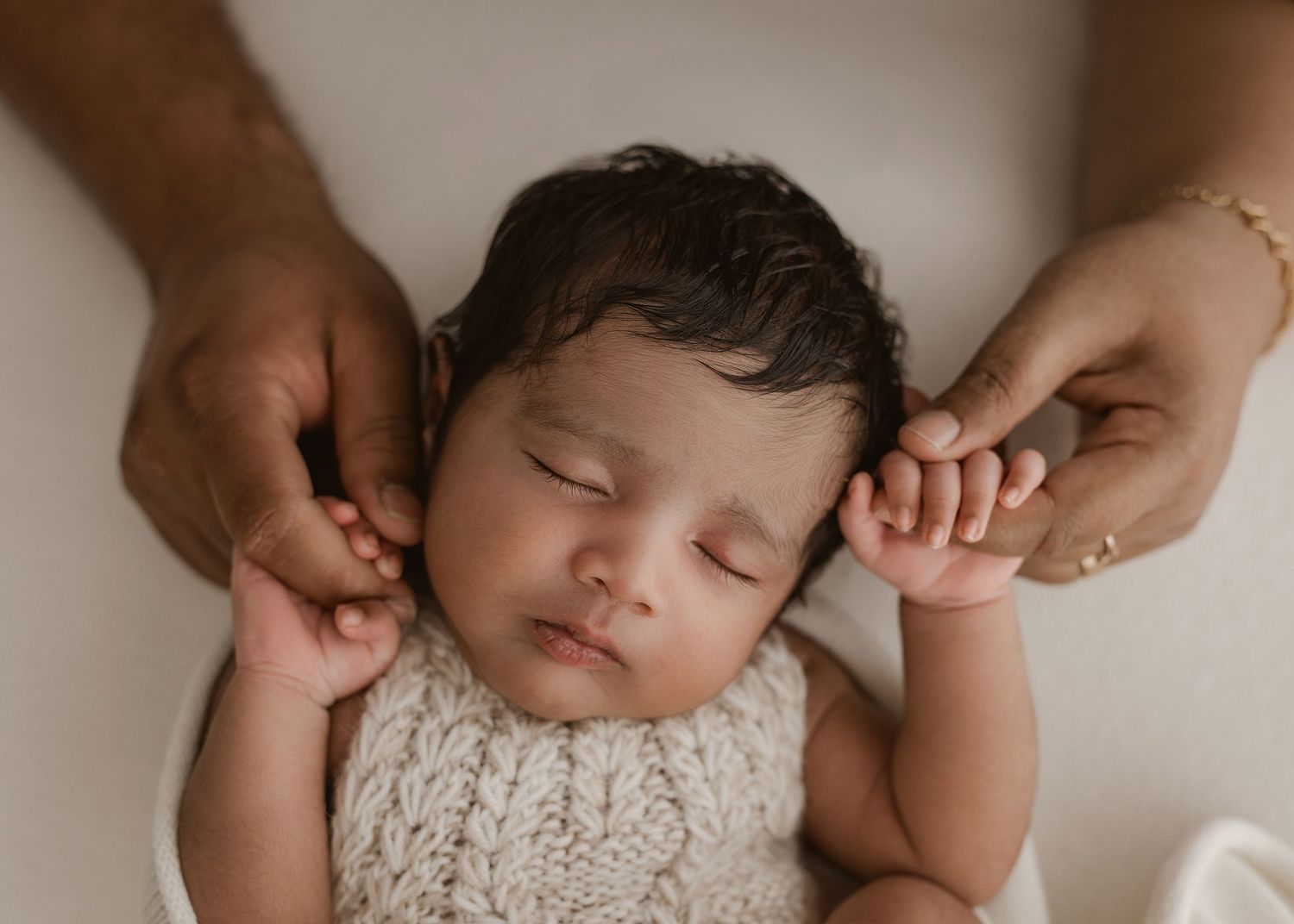 Sleeping baby in knitted white outfit being gently held by adult hands.