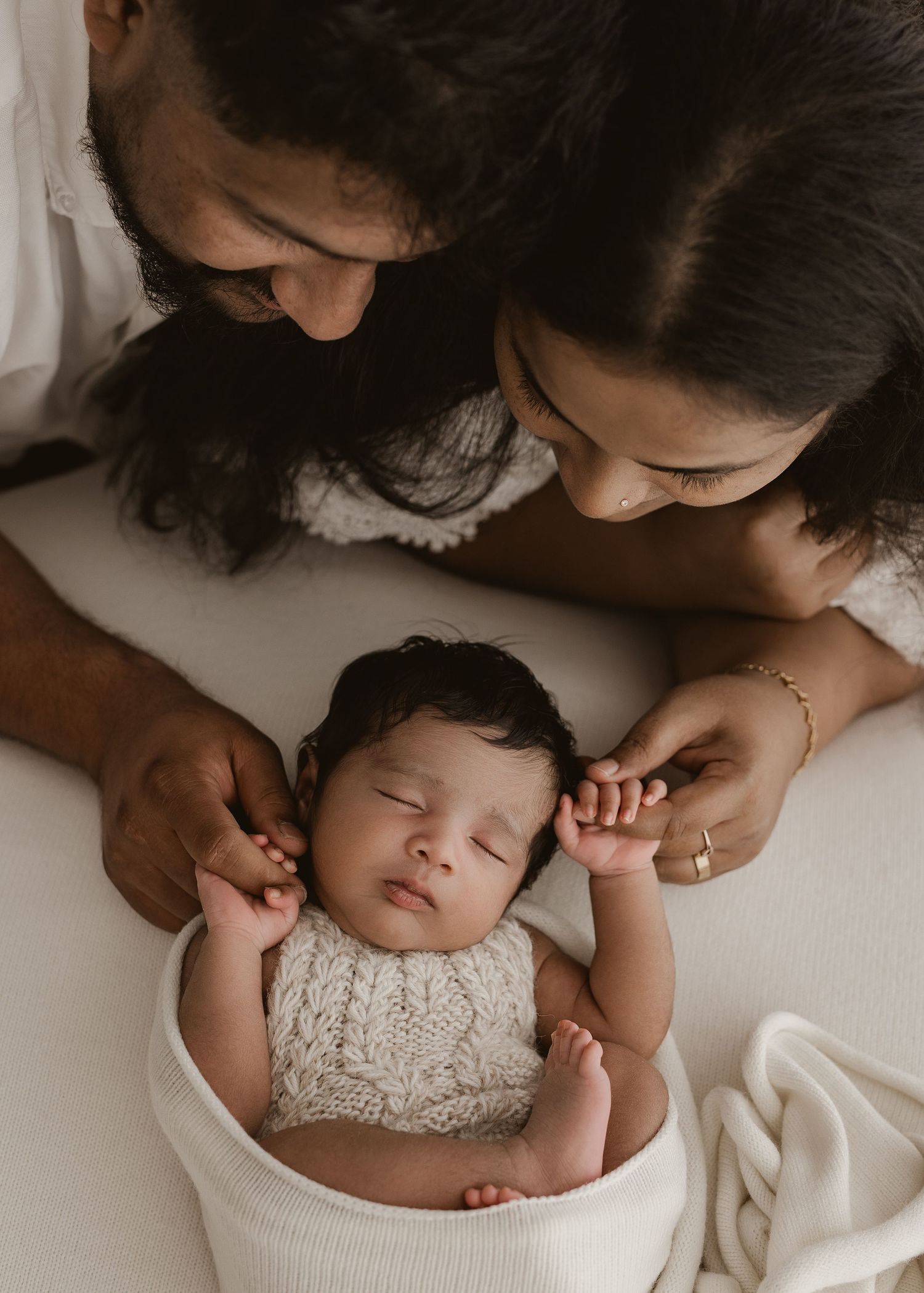 A sleeping baby in a white lace dress lies peacefully in a basket while two people lovingly watch from above.