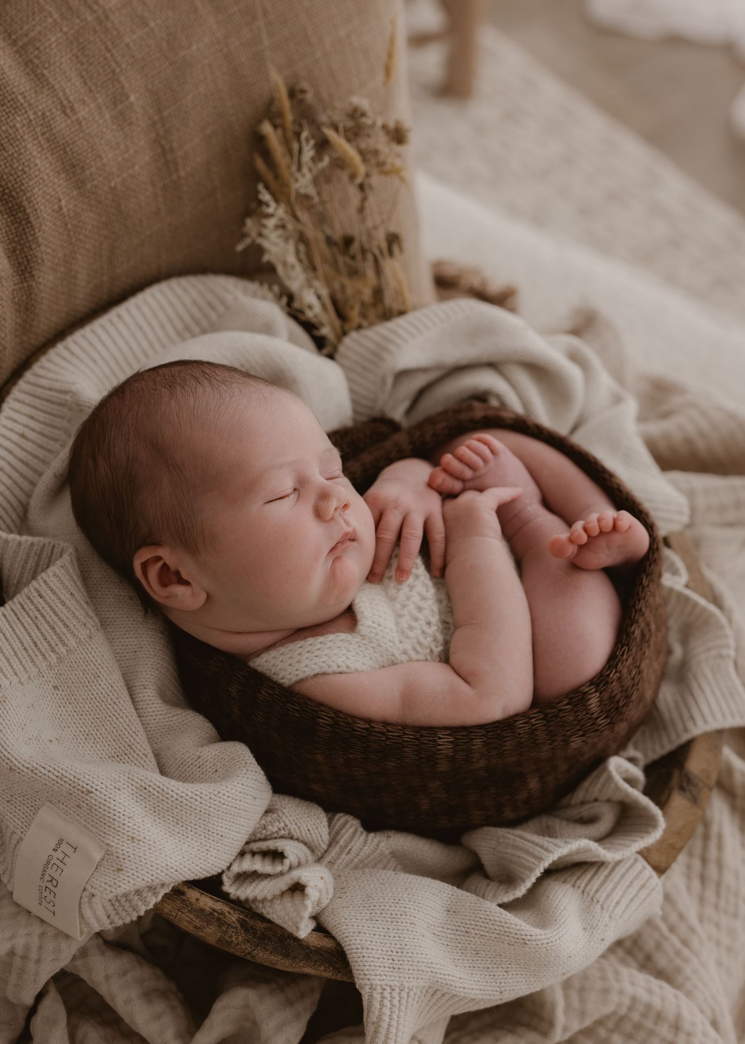 A sleeping newborn baby lies peacefully on a cream-colored knit blanket in a rustic brown basket.