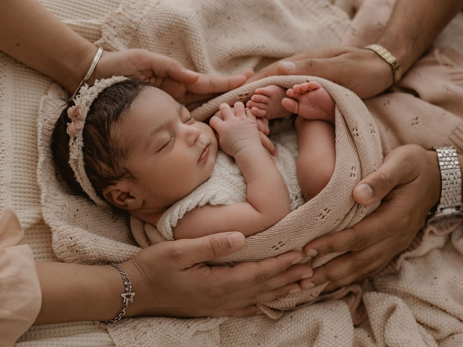A sleeping newborn baby wearing a white lace headband lies cradled in gentle hands on soft beige fabric.