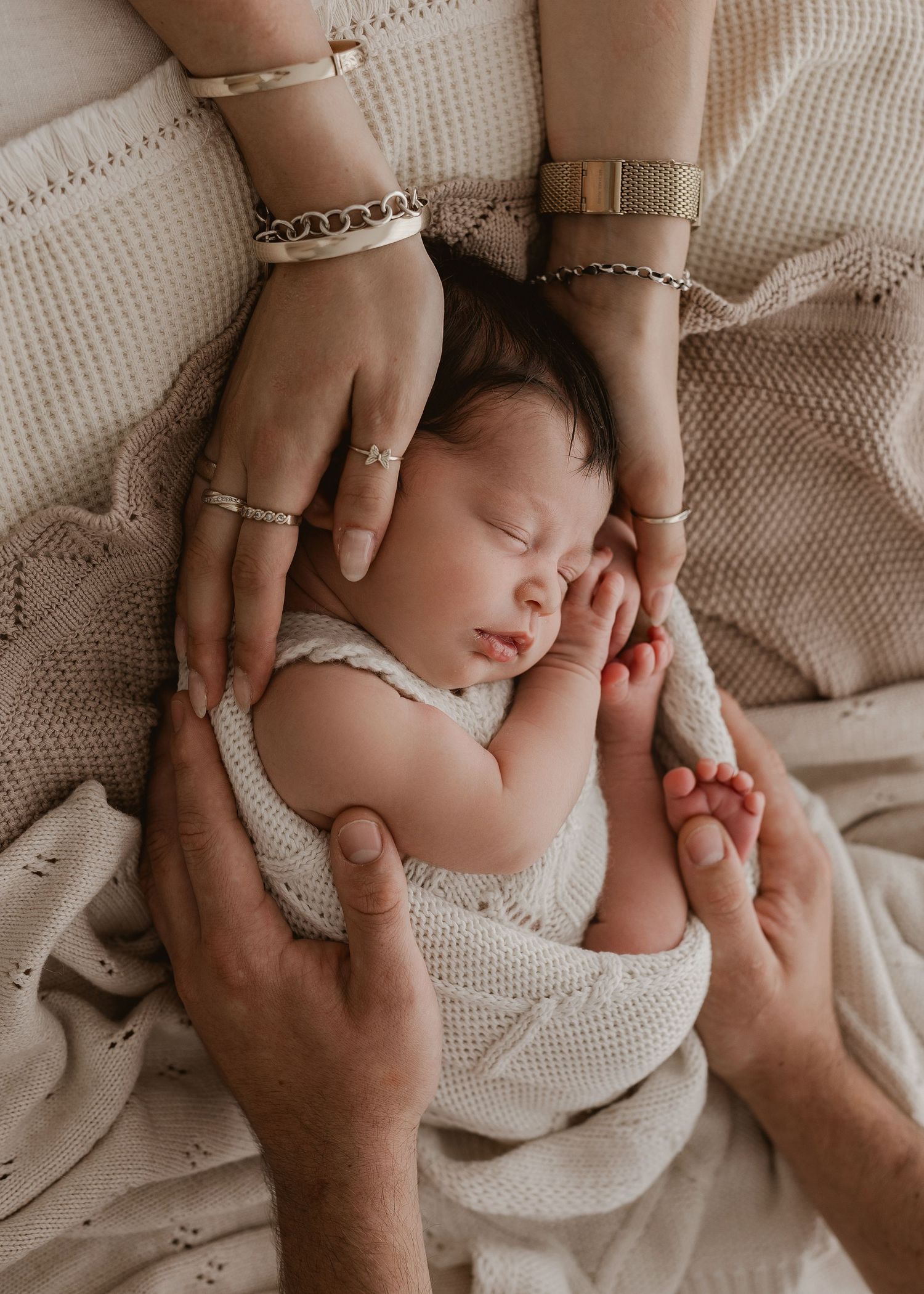 Gentle hands tenderly caress a sleeping newborn baby wrapped in soft white knit blanket.