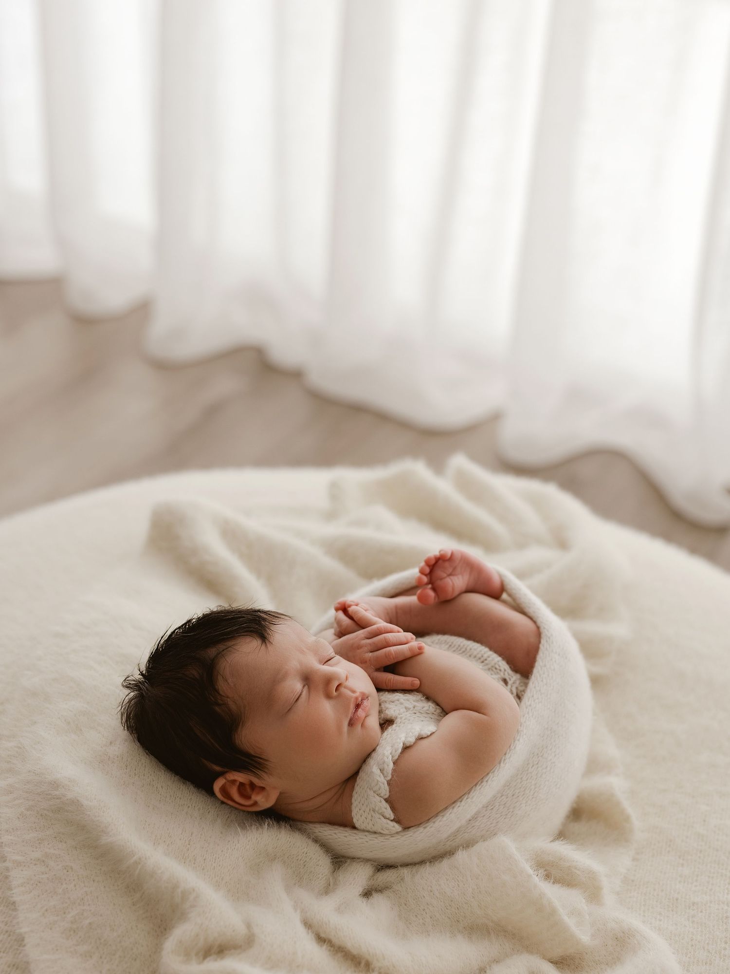 Peaceful sleeping baby wrapped in soft white blanket lying on cream colored surface near sheer curtains.