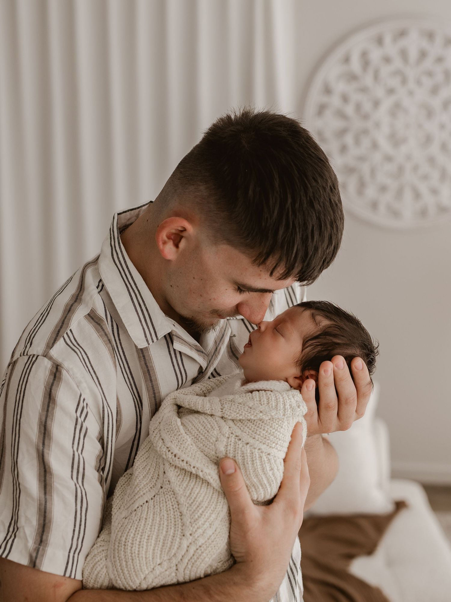 Parent in striped shirt lovingly embraces and kisses sleeping infant in white knit sweater.