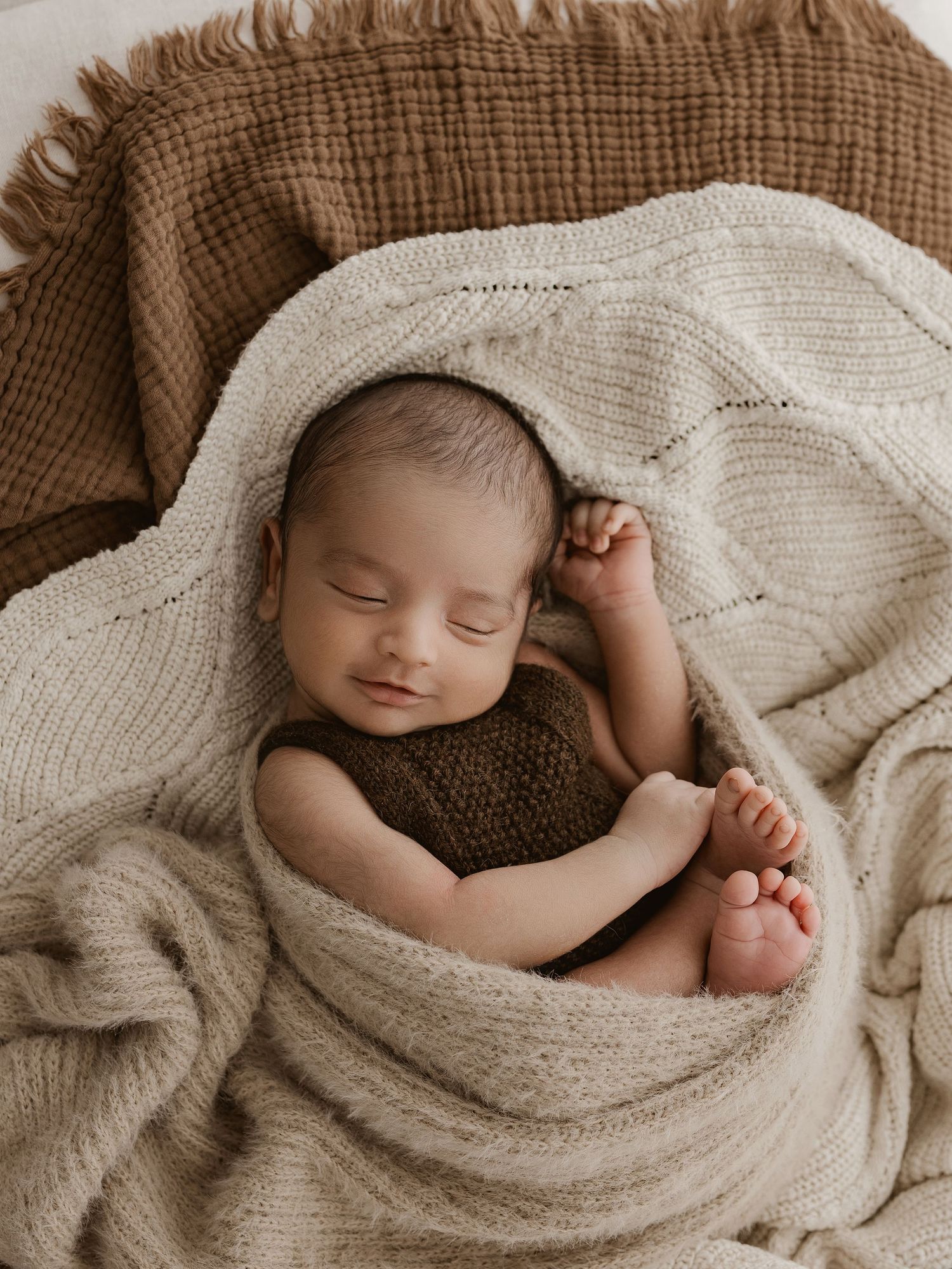 Peaceful sleeping newborn wrapped in cozy beige and brown knit blankets on textured fabric background.