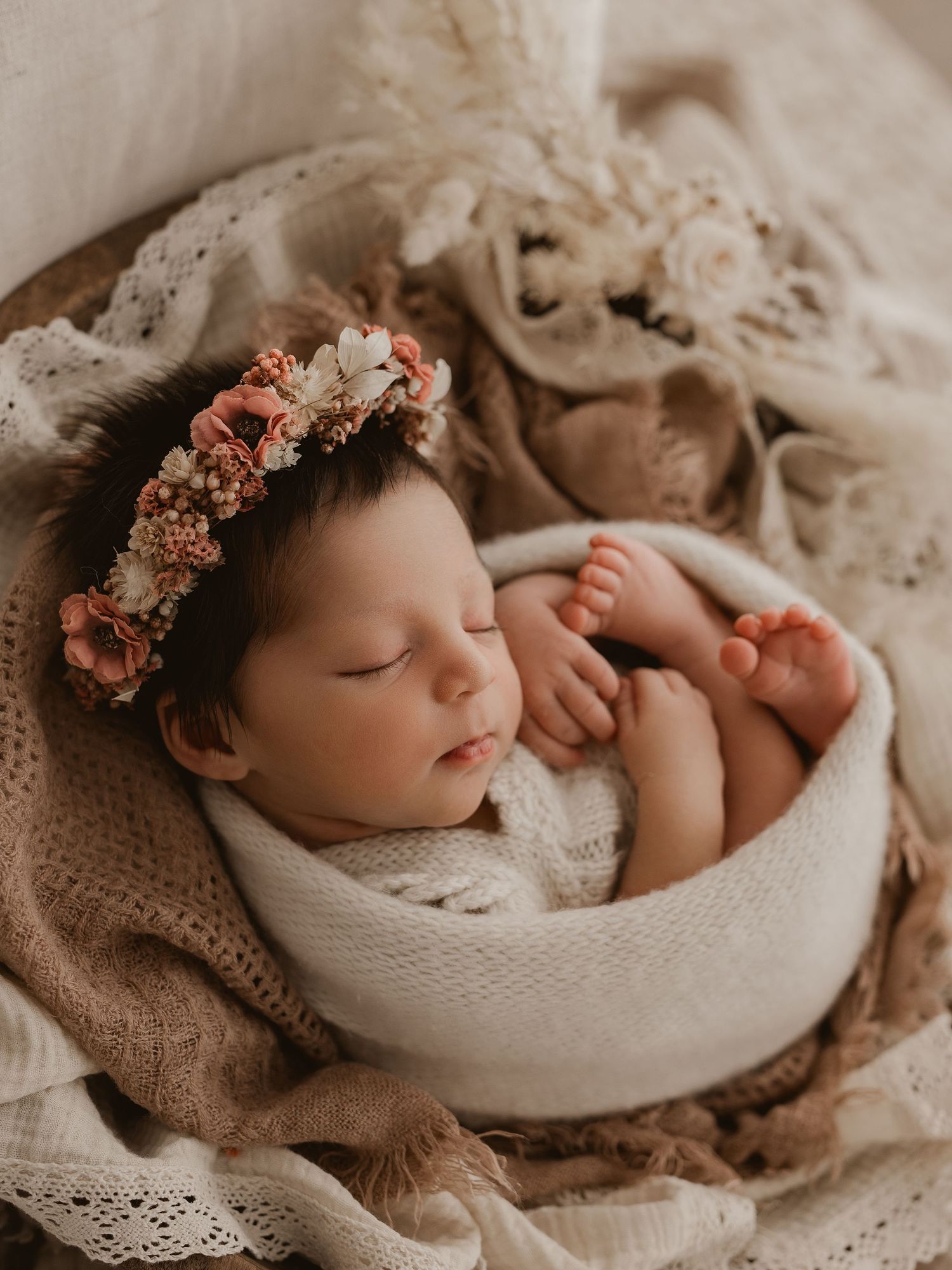 Peaceful newborn sleeping in soft cream-colored bedding with delicate floral crown.