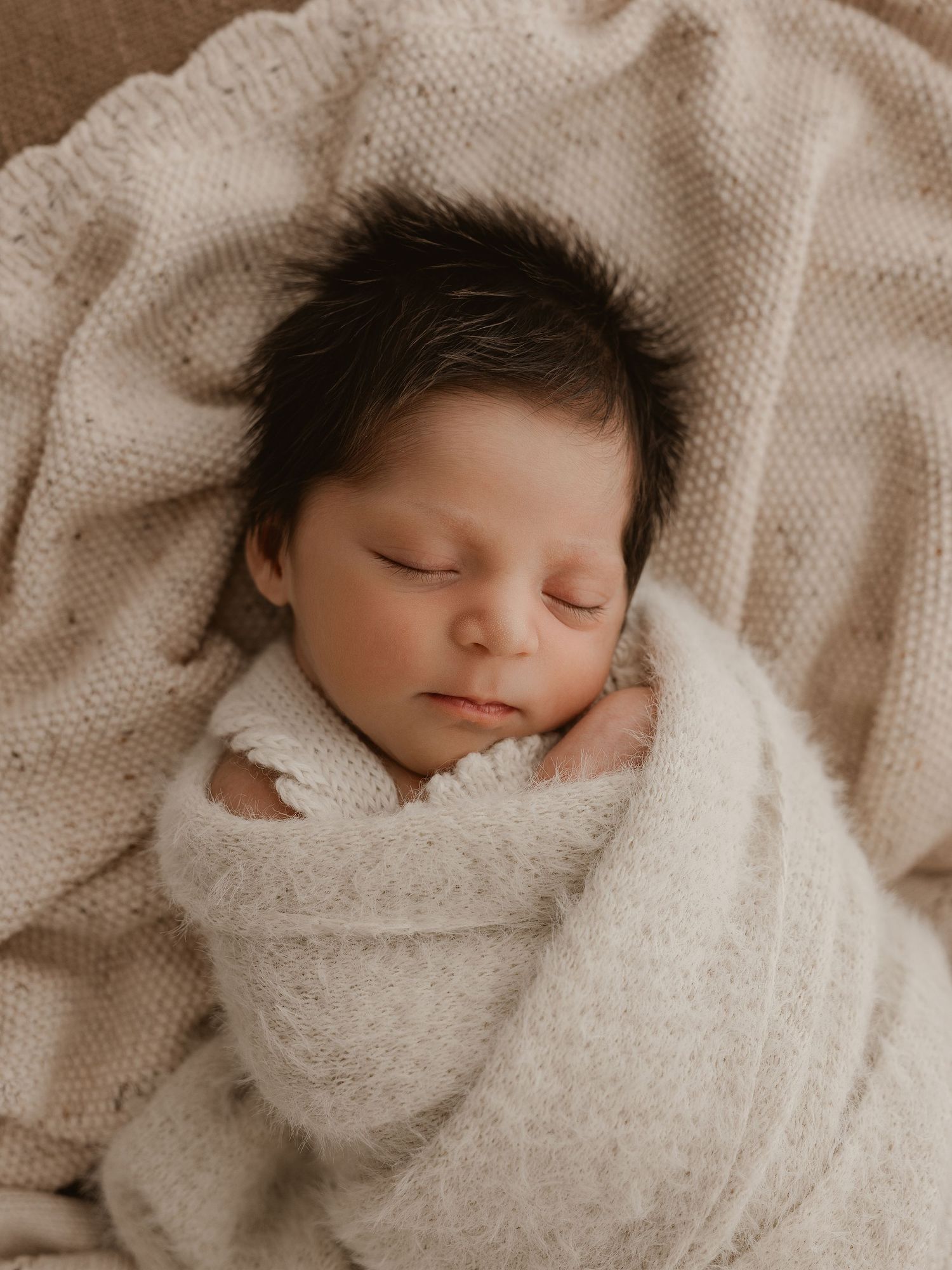 Sleeping newborn baby wrapped in cozy white knit blanket against neutral beige background.