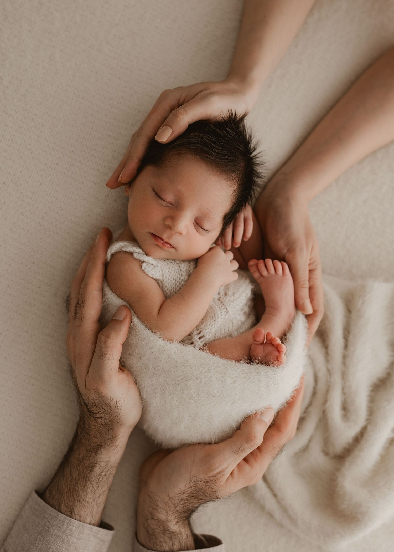 A newborn baby rests peacefully on a soft white blanket while gentle hands provide comfort and support.