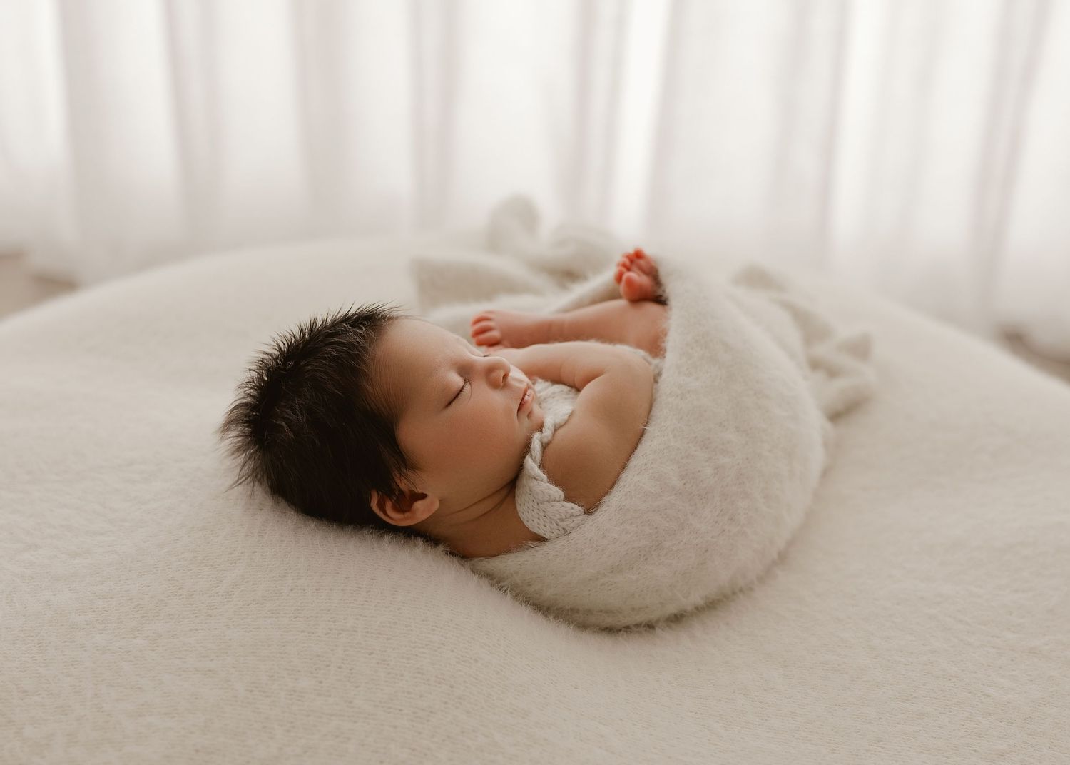 A sleeping baby swaddled in white fabric lies on a cozy white blanket in a peaceful setting.