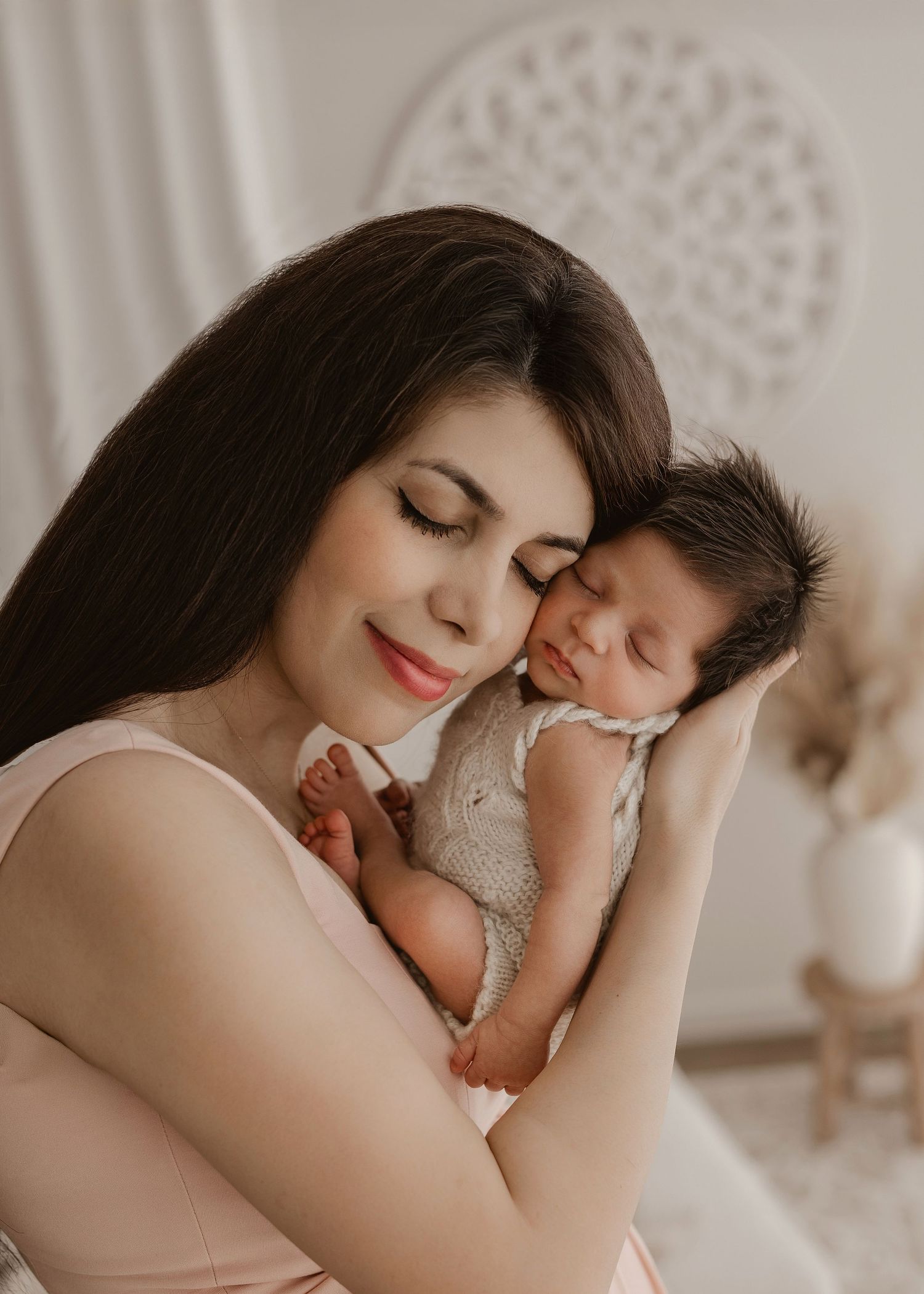 A tender moment between mother and newborn baby in soft neutral tones against a white macrame backdrop.