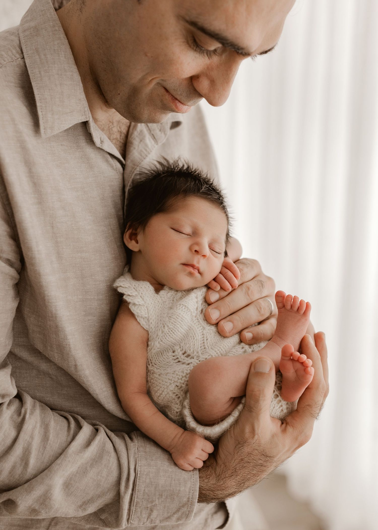 A tender moment as someone holds a sleeping newborn baby wearing white lace against a light background.