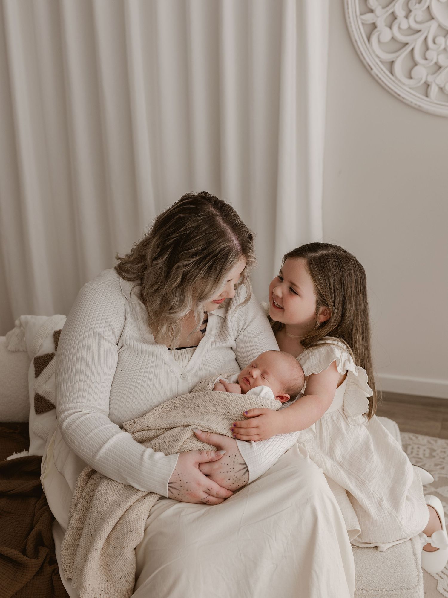 A tender family moment captured in soft neutral tones as they cuddle with a newborn baby on a white chair.