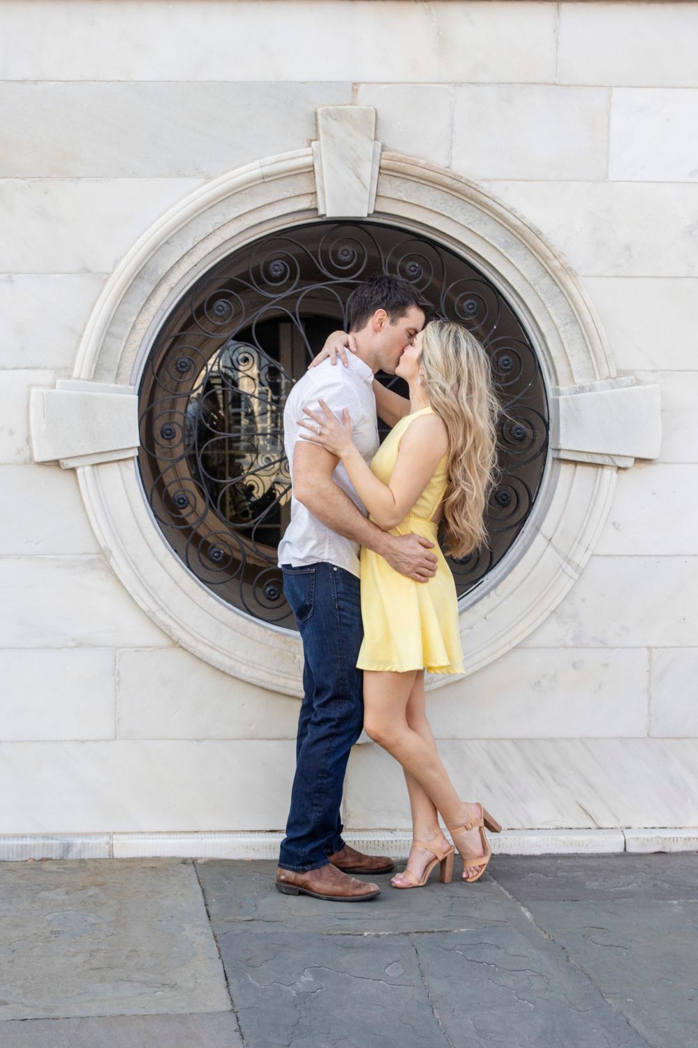 Couple sharing intimate moment in front of ornate circular window with wrought iron details.