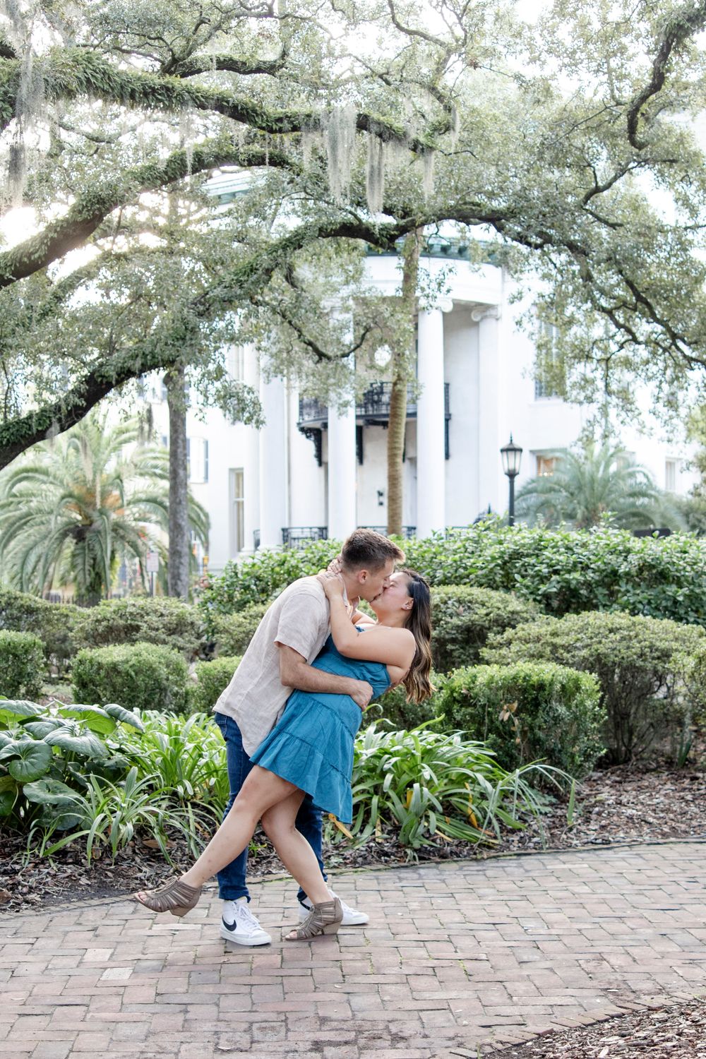 Romantic couple embraces on brick path under moss-covered trees near white building.