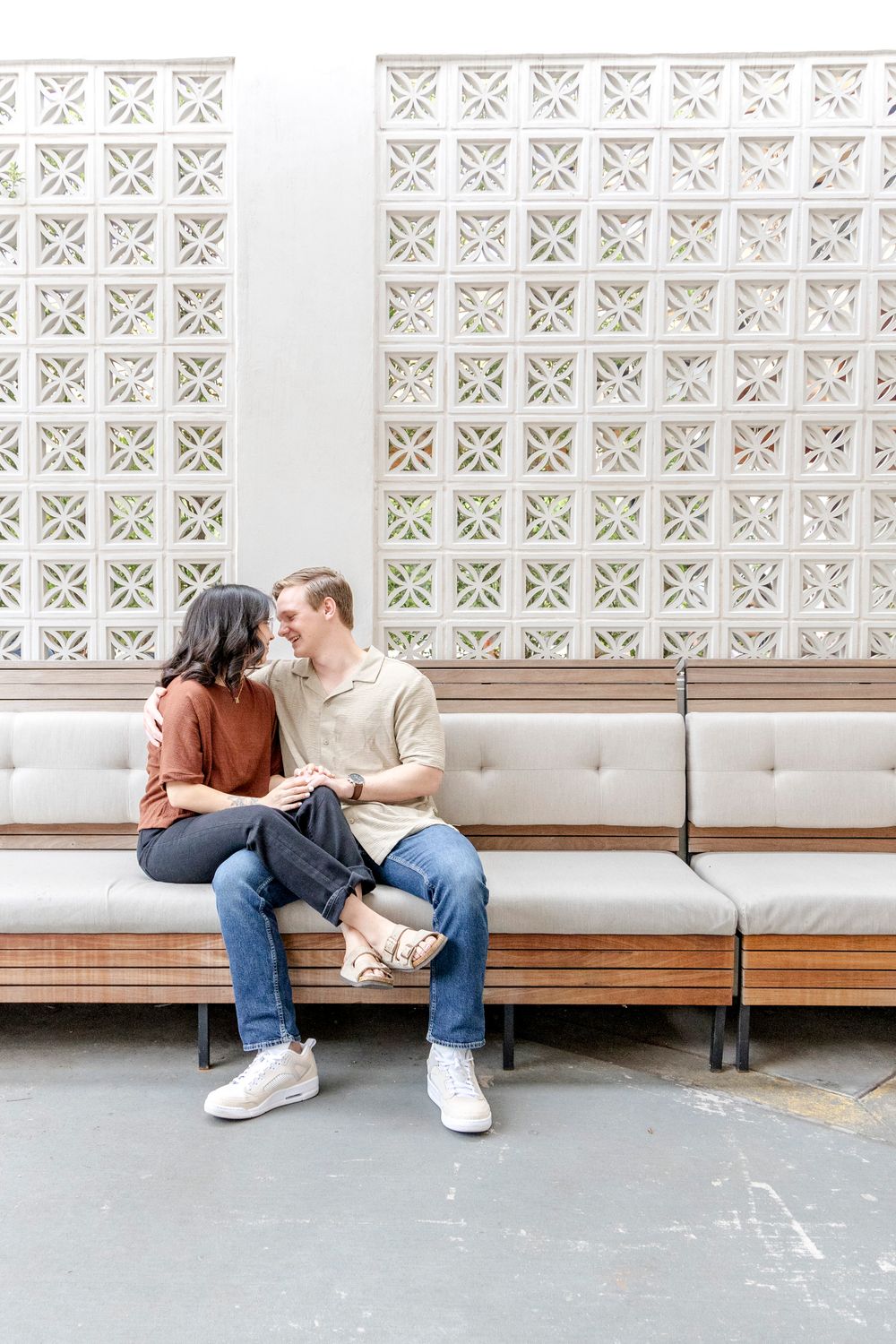 Two people in casual outfits sit close together on a bench against a decorative white lattice wall pattern.