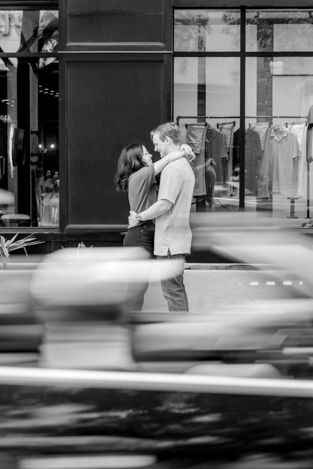 A romantic black and white moment captured outside a storefront with motion blur of passing cars in the foreground.