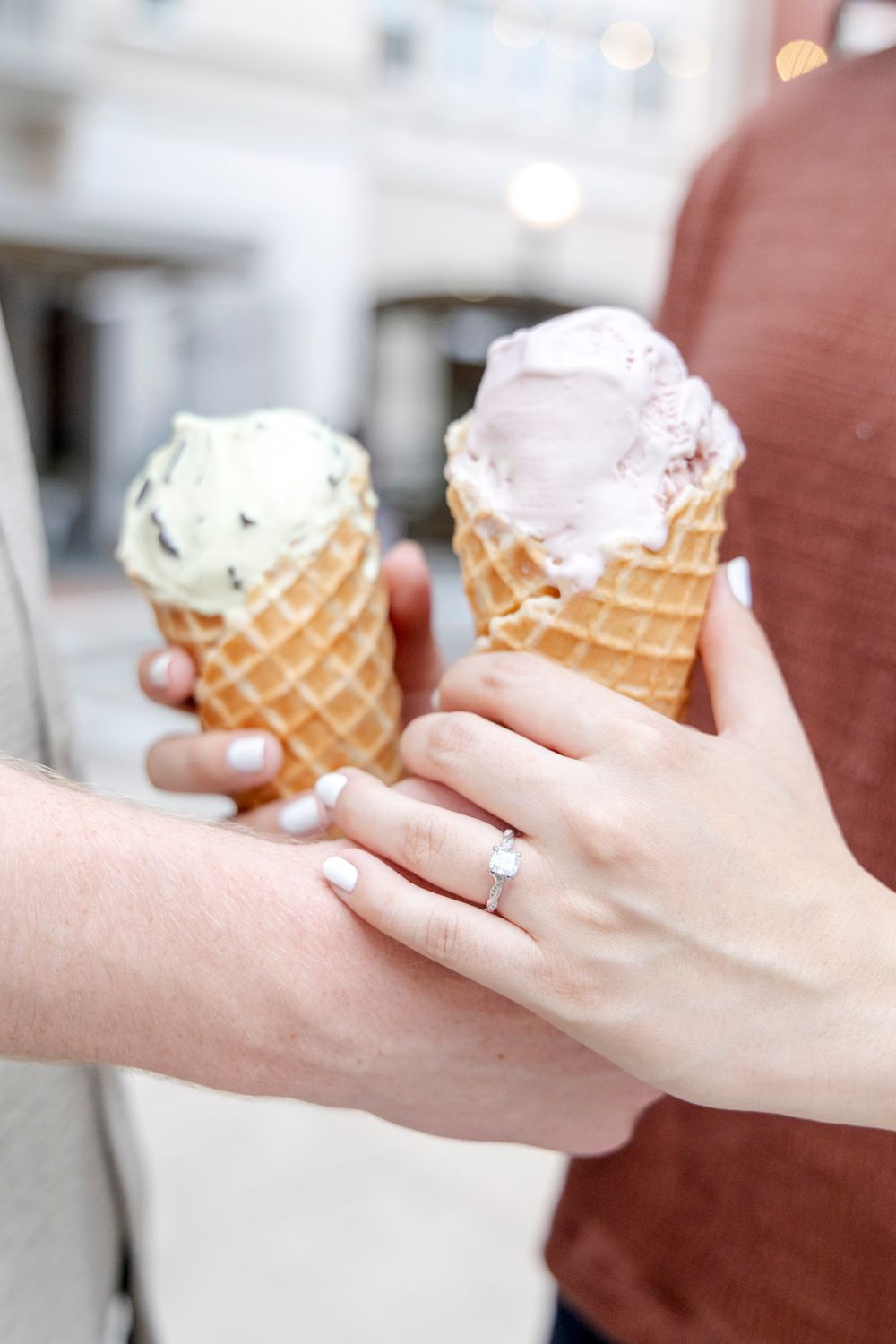 Two hands hold ice cream cones with pink and white scoops in waffle cones as one person shows off their engagement ring.