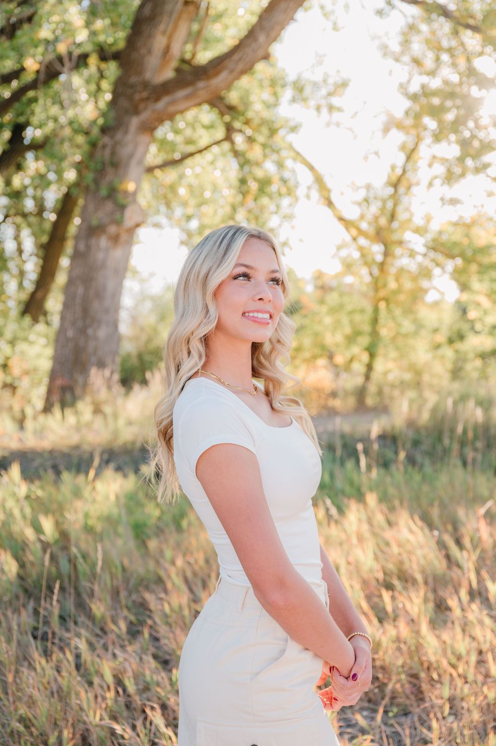 Outdoor portrait in a white dress against sunlit trees and natural grass landscape.