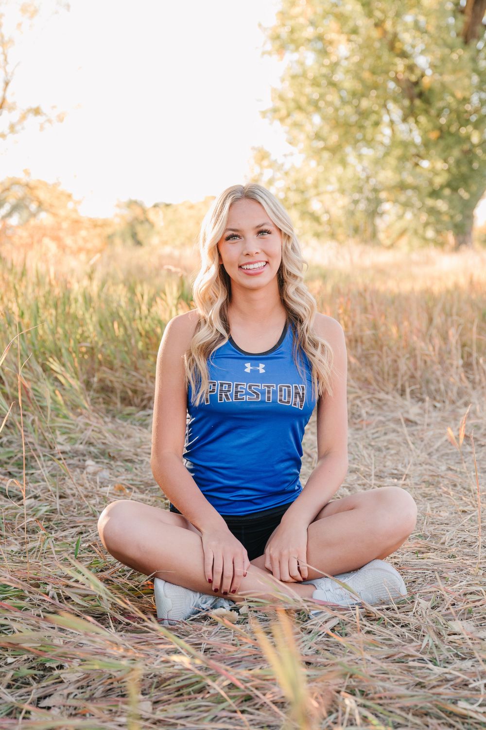 Athletic portrait in blue tank top sitting cross-legged in golden field at sunset.