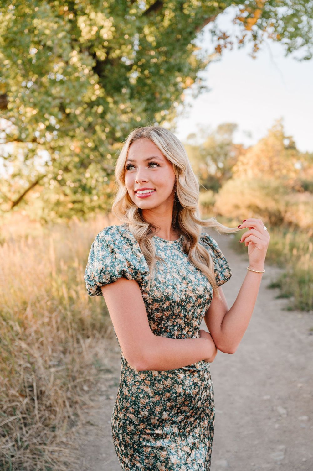 Someone in a floral midi dress poses on a dirt path during golden hour with trees and warm sunlight in the background.
