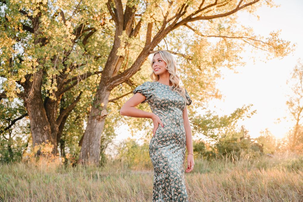 In a sunlit meadow, a figure in a floral dress twirls gracefully beneath golden-lit trees at sunset.