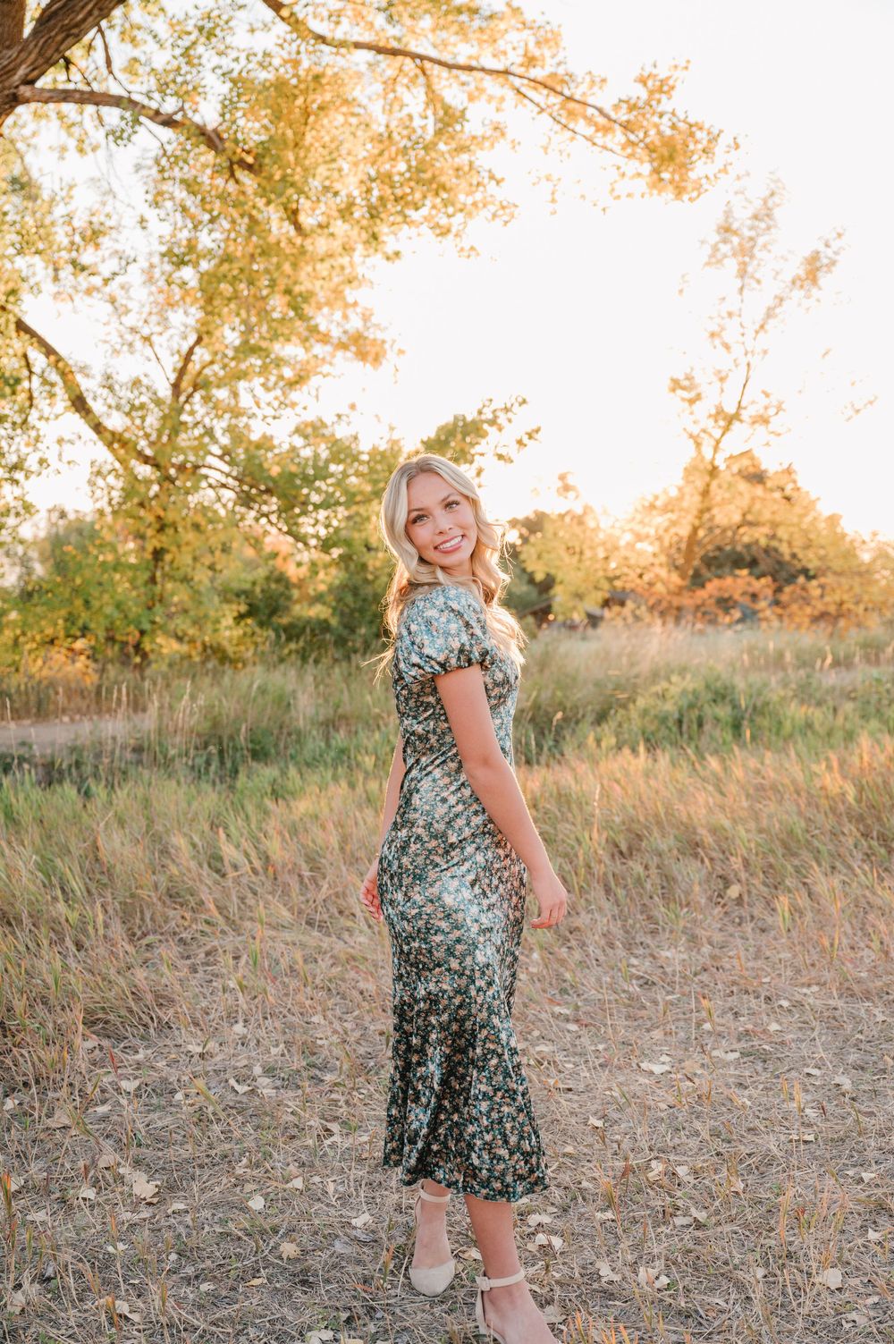 A woman in a floral dress poses in a sun-lit field with golden tall grass and autumn trees in the background.