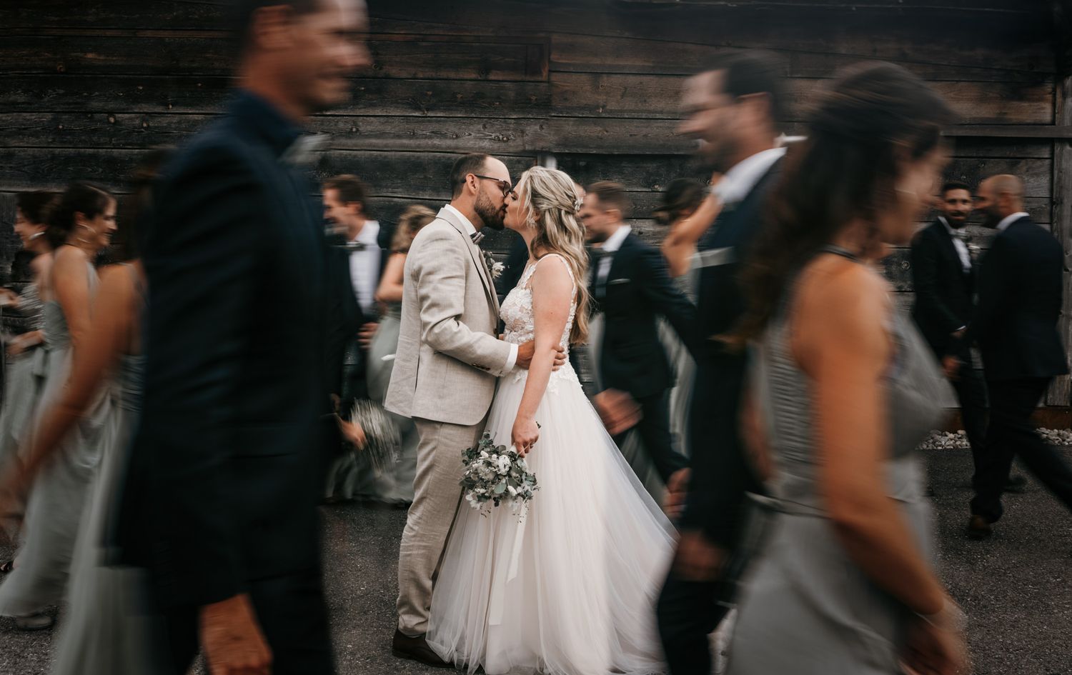 Bride and groom share a kiss surrounded by blurred wedding guests in motion at their reception.
