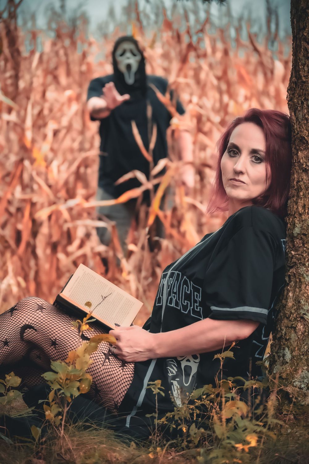 Woman reading a book outdoors near cornfield with Ghostface from Scream lurking behind her in Halloween costume.