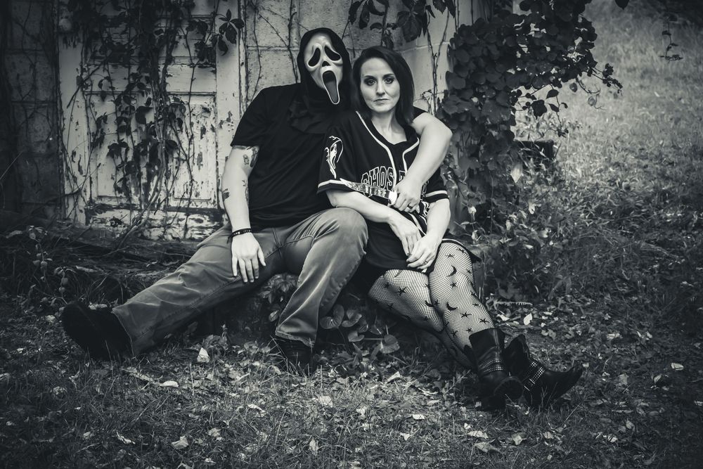 Black and white photo of couple sitting outdoors, one wearing Ghostface mask, against rustic wall with ivy.